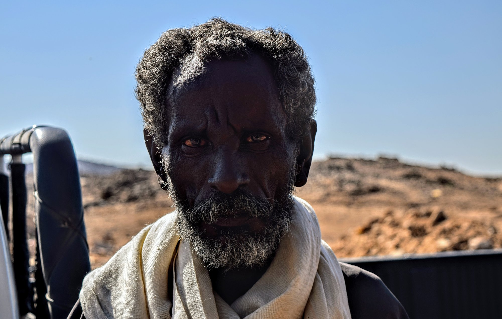 L'image montre un homme avec une peau foncée et des cheveux crépus, portant une écharpe claire autour du cou. Son regard est intense et semble exprimer une certaine sagesse ou expérience. L'arrière-plan est flou et semble représenter un paysage désertique avec des montagnes ou des collines à distance. La lumière est vive, ce qui met en valeur les traits du visage de l'homme.