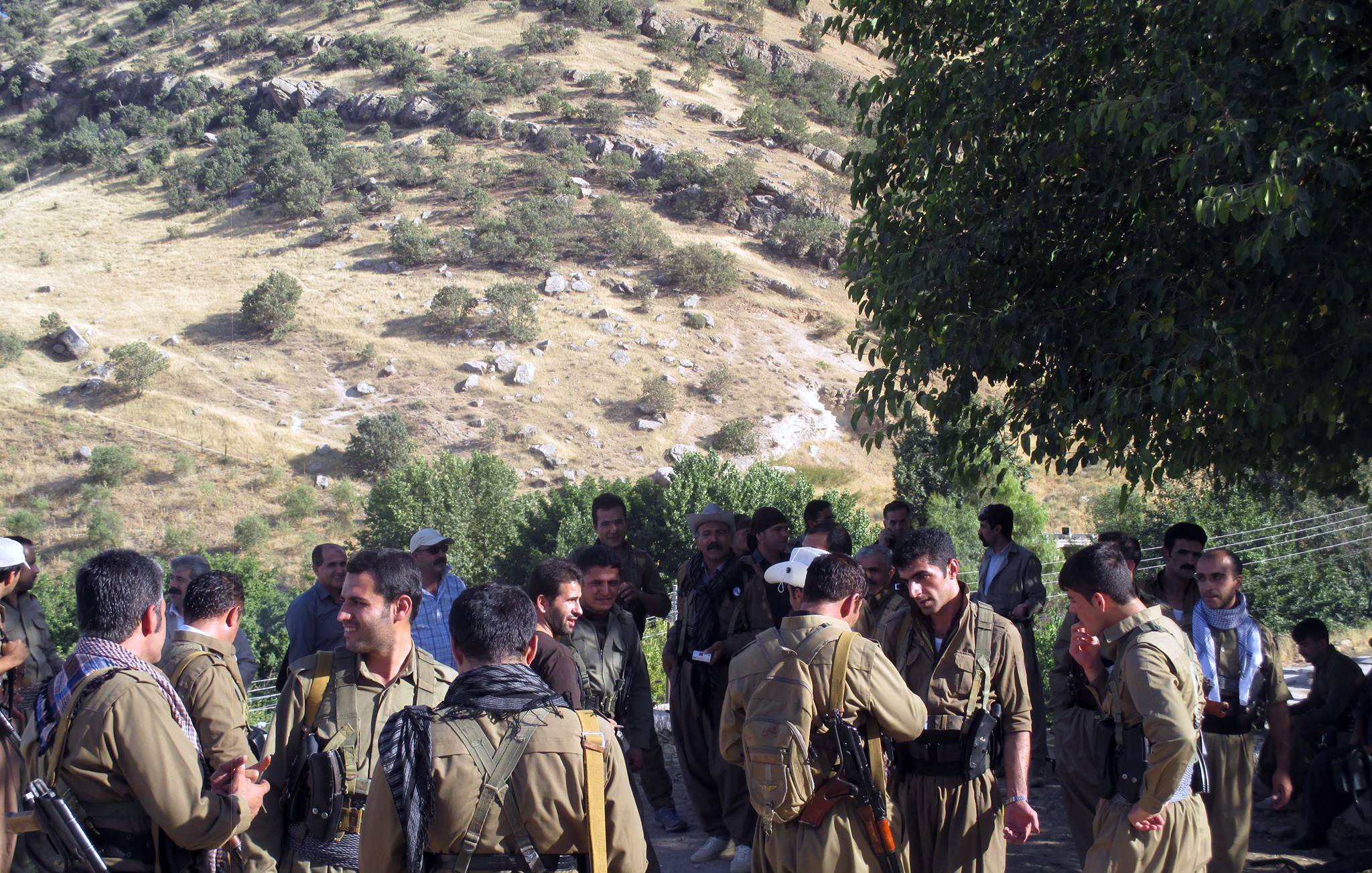 الصورة تظهر مجموعة من الرجال يرتدون ملابس عسكرية في منطقة طبيعية. توجد تلال خضراء في الخلفية، وبدت بعض الأشجار. يبدو أن هؤلاء الرجال يتفاعلون مع بعضهم البعض، حيث هناك من يتحدث ومن يتواجد في مجموعات صغيرة. يظهر في الصورة أيضا بعض الأشخاص يحملون أسلحة. الجو العام يوحي بنشاط اجتماعي أو تحضير لشيء ما.