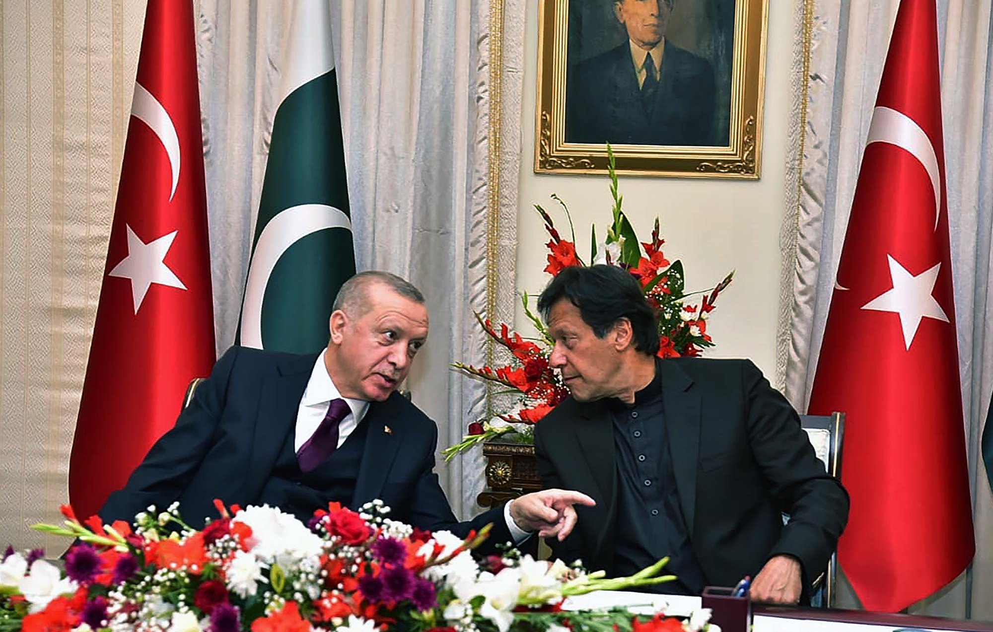 The image features two men engaged in conversation, likely discussing matters of significance. They are seated at a table adorned with flowers, and the backdrop includes flags of Turkey and Pakistan prominently displayed. A portrait of a historical figure hangs on the wall above them. Both individuals appear to be in formal attire, suggesting an official or diplomatic context for their meeting.