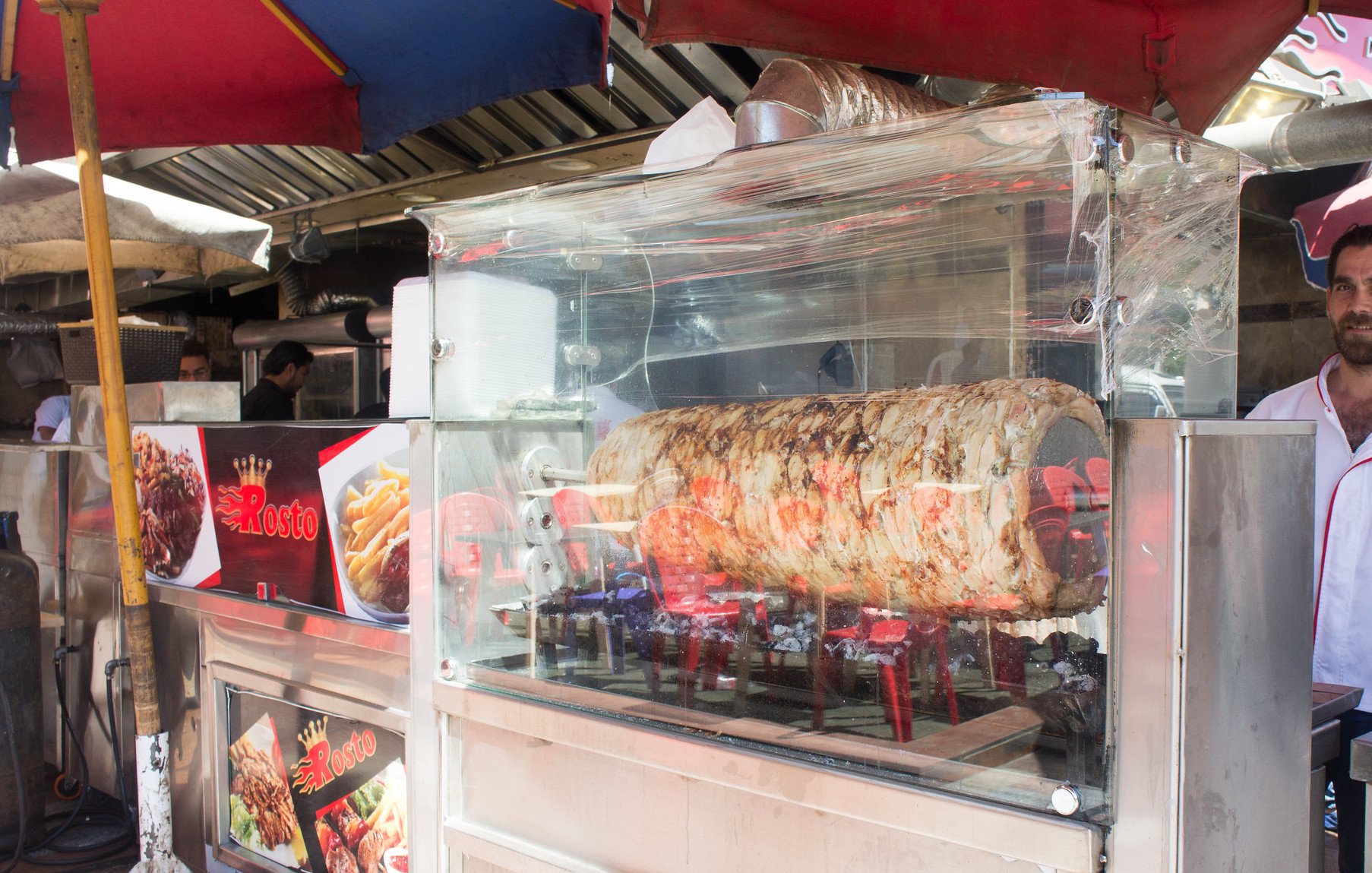 The image depicts a food stall featuring a large kebab or shawarma that's roasting on a vertical spit. The meat is visibly browning as it cooks, and there are flames underneath. The stall is covered by colorful umbrellas providing shade. In the foreground, a man stands behind the counter, likely the vendor, appearing to interact with customers or prepare food. The stall also displays menu items on signs, emphasizing its offerings. The atmosphere looks lively, typical of a street food setting.