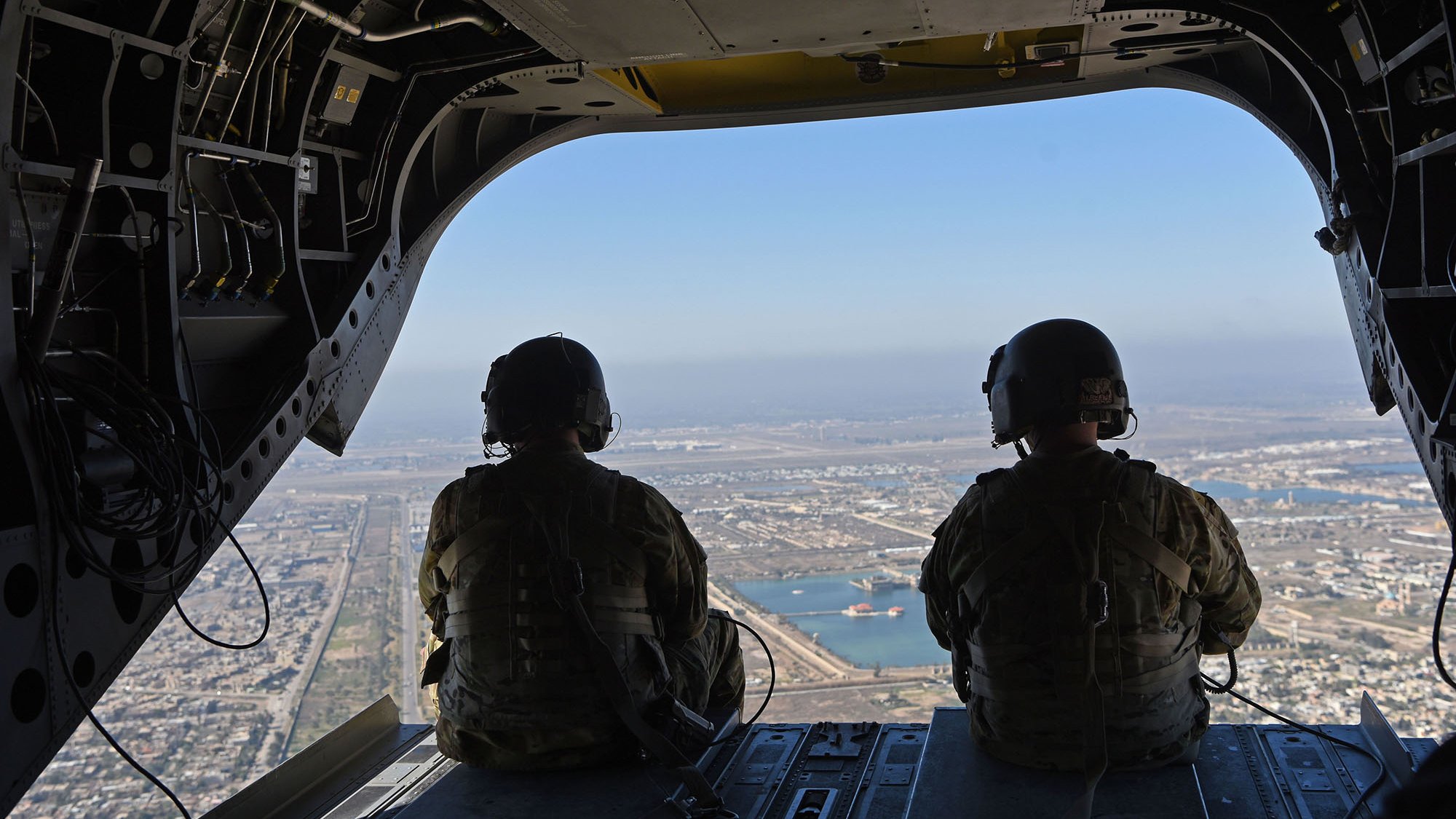 تظهر الصورة مشهداً من داخل طائرة عسكرية، حيث يجلس جنديان يرتديان خوذات وسترات عسكرية. يتم تصويرهما من الخلف وهما يتطلعبان من فتحة الطائرة. يظهر في الأفق منظر للمدينة مع ممرات للمطار وبعض المباني والماء في الخلفية. السماء واضحة وتبدو هادئة.