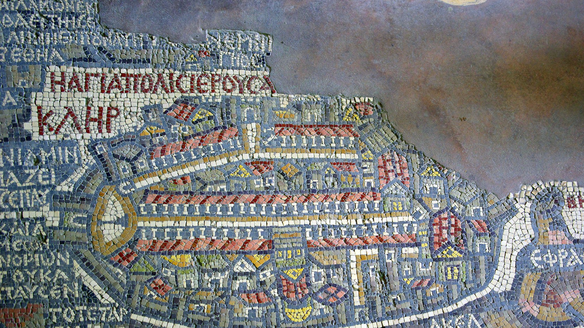 الصورة تظهر فسيفساء تاريخية تمثل خريطة لمكان ما، حيث تحتوي على تفاصيل معمارية وشوارع، بالإضافة إلى نصوص مكتوبة باللغة اليونانية. يظهر فيها تصميم معقد يوضح ملامح المدينة، بما في ذلك الأبنية والمنافذ الرئيسية. الألوان المستخدمة تشمل الأحمر والأزرق والأبيض، مما يضفي جمالية على العمل الفني. هذه الخريطة تعكس التاريخ والثقافة المعمارية للمنطقة التي تمثلها.
