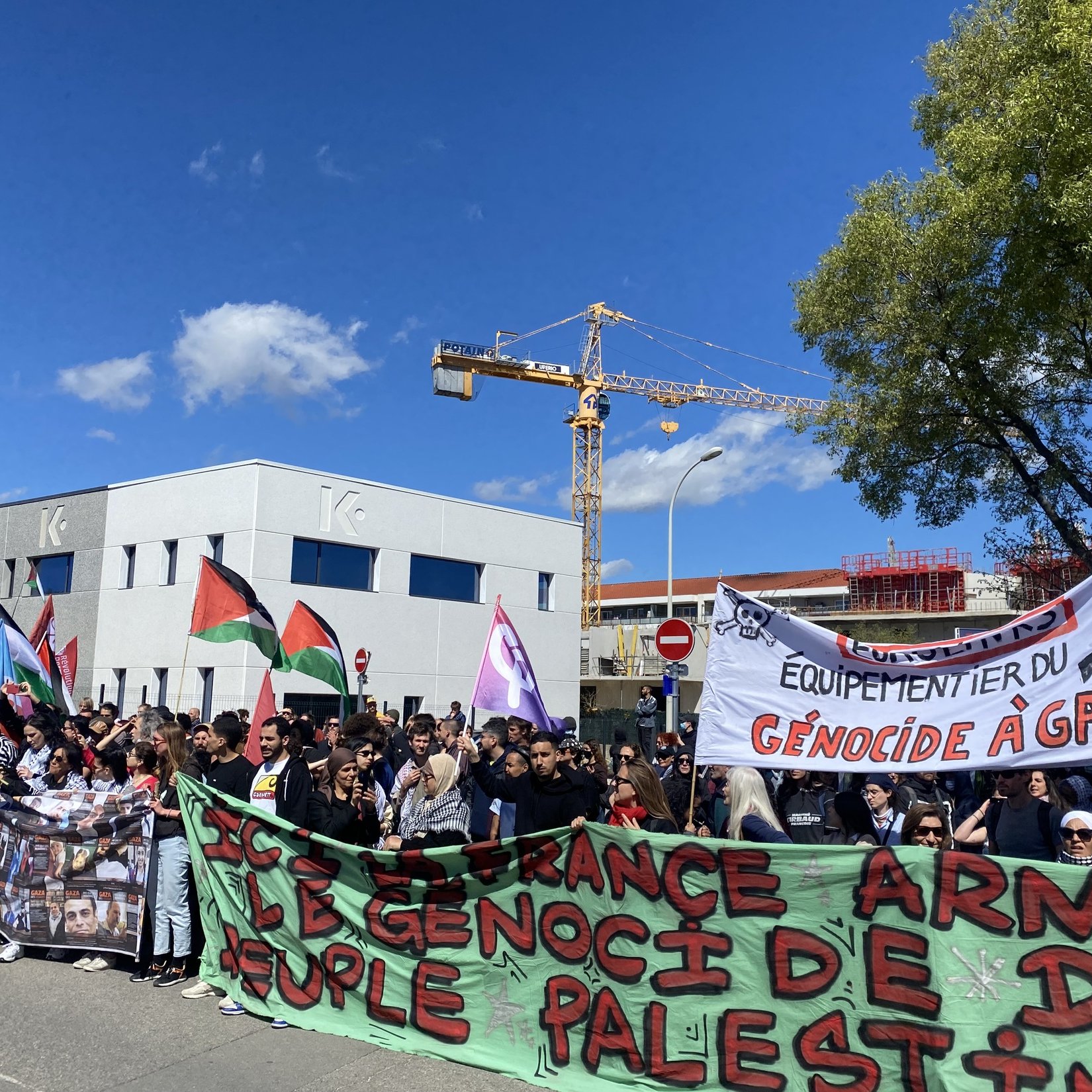 L'image montre une manifestation avec une grande foule de personnes rassemblées. Les manifestants portent des bannières et des drapeaux, y compris des drapeaux palestiniens. Certains panneaux affichent des messages en faveur des droits du peuple palestinien et dénoncent le génocide à Gaza. L'ambiance semble être celle d'une protestation pacifique, avec des participants unis pour une cause commune. En arrière-plan, on peut voir des bâtiments et une grue de construction.