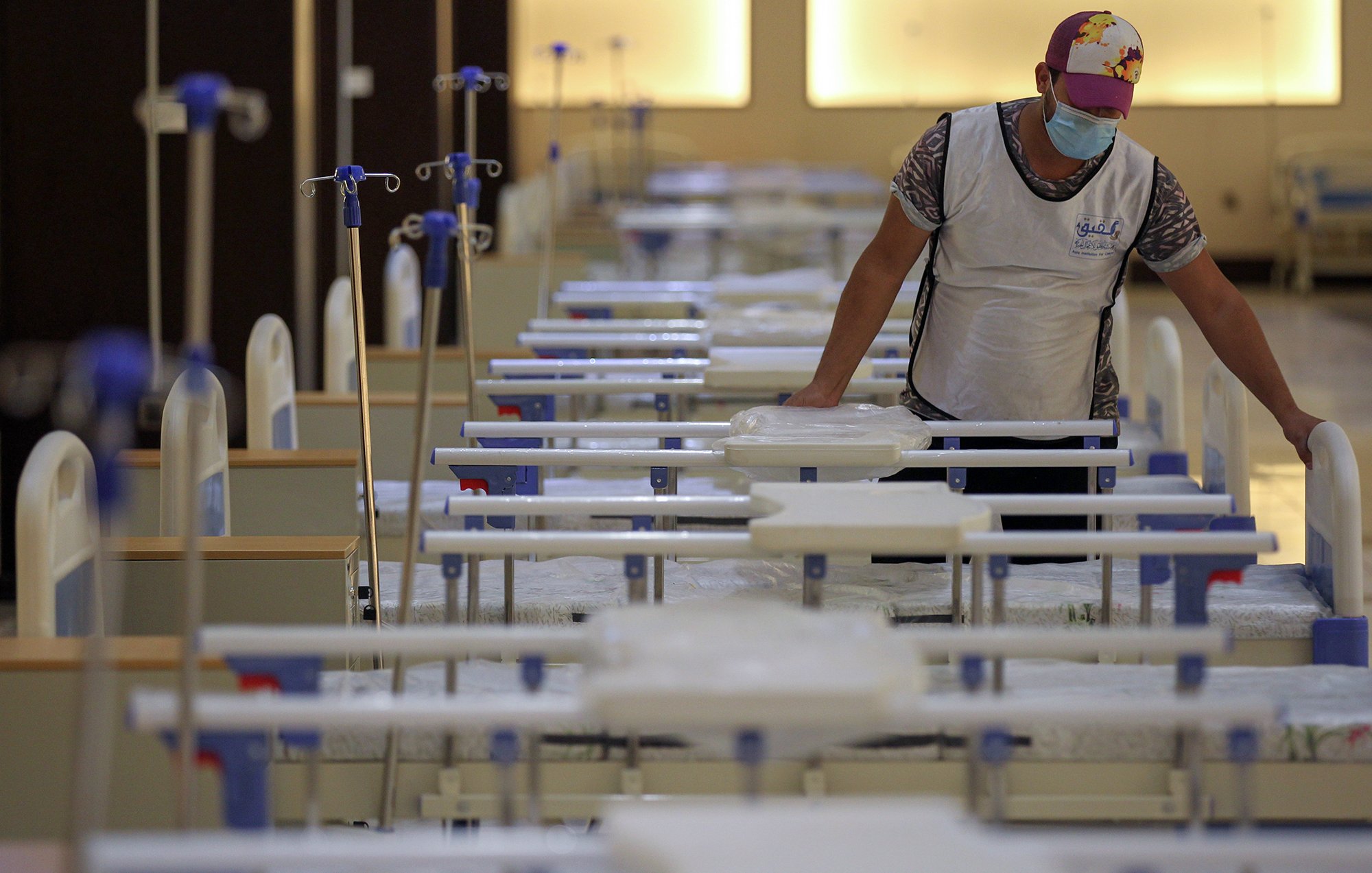 ترتدي الصورة مشهداً داخل مرفق صحي، حيث يظهر شخص واحد يرتدي كمامة وقميصاً. الشخص يركز على سرير طبي بين عدة أسرّة مرتبة بشكل منظم في غرفة واسعة مضاءة بشكل جيد. تظهر في الخلفية أعمدة ومعدات طبية، مما يوحي بأن المكان يُستخدم لعلاج المرضى. يتميز الجو بالهدوء والترتيب.