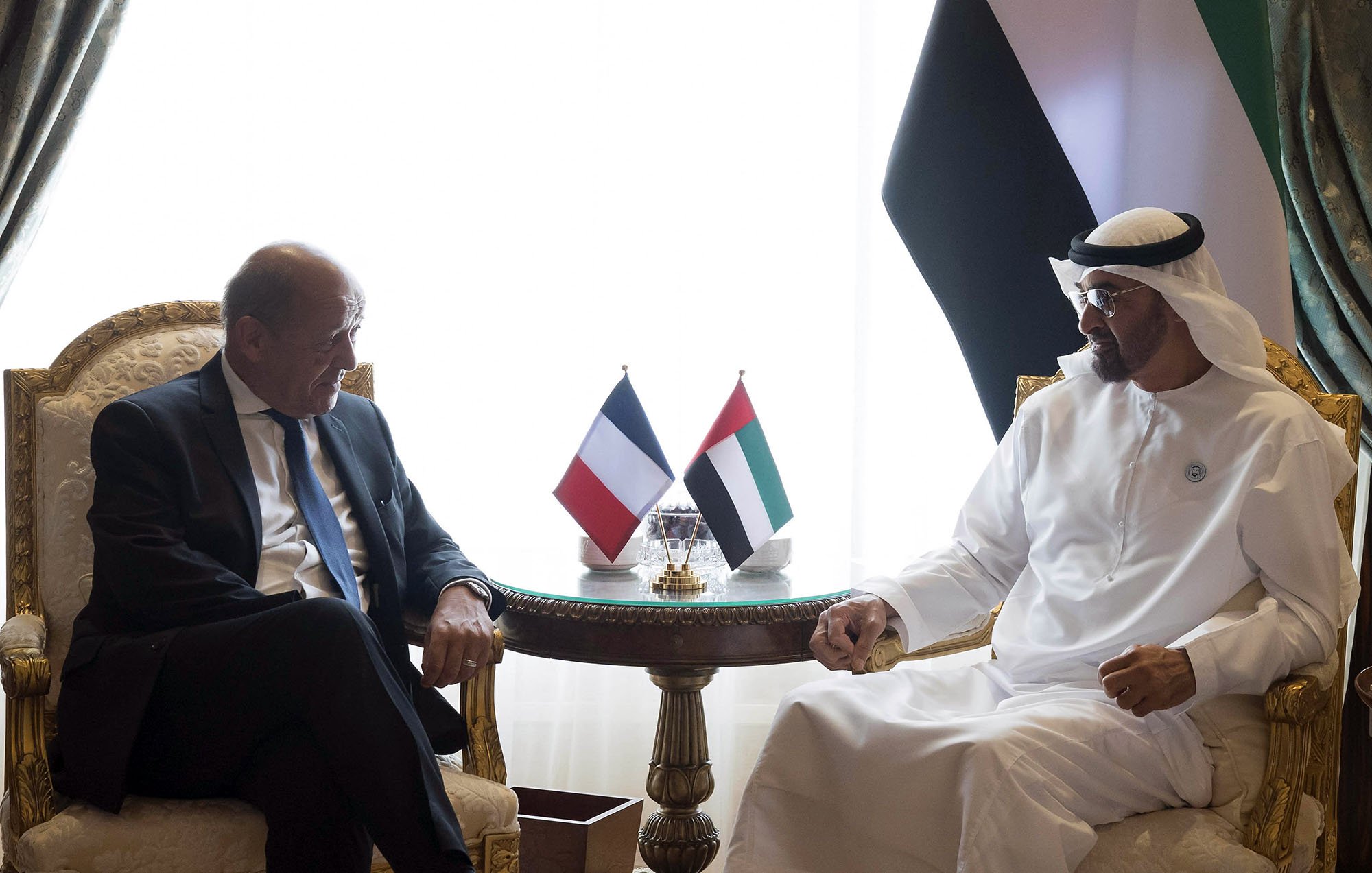 The image shows two men seated at a table in a luxurious setting. They are engaged in conversation, appearing to be in a diplomatic or formal meeting. There are flags on the table representing France and the United Arab Emirates. The background features elegant curtains and decor. One man is dressed in a suit, while the other is wearing traditional Emirati attire.