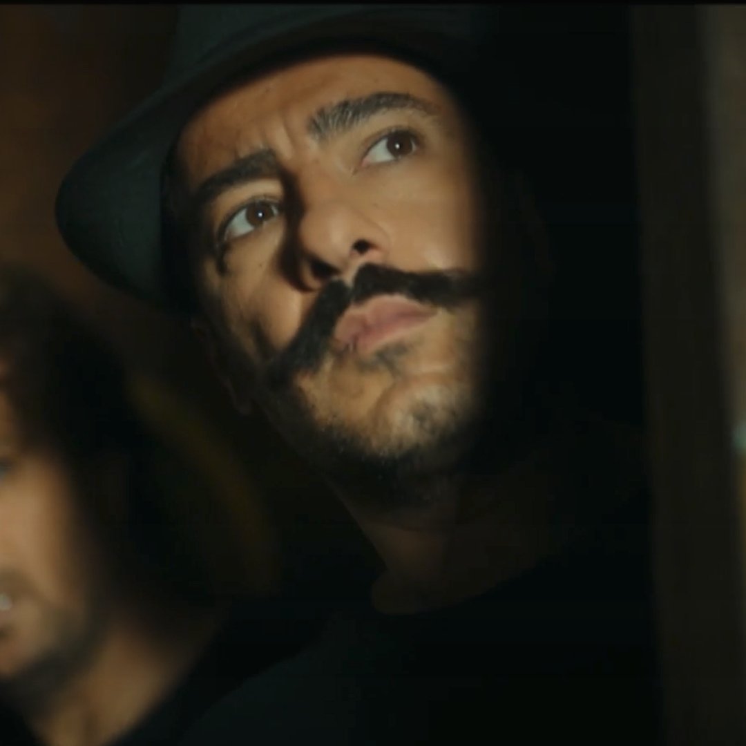 در این تصویر، دو مرد در یک محیط تاریک و مرموز دیده می‌شوند. مردی در جلو دارای کلاهی سیاه و سبیلی خاص است که به جلو نگاه می‌کند و حالت تفکر یا نگرانی دارد. مرد دیگر در پس‌زمینه، با ظاهری نگران و معتمد، از سمت چپ او به تصویر نگاه می‌کند. نور به صورت جزئی روی صورت‌های آن‌ها می‌تابد و حس تنش یا خطر در صحنه وجود دارد.