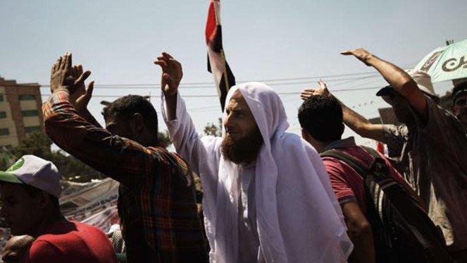 L'image montre une scène de rassemblement ou de manifestation. On peut voir un groupe de personnes levant les bras, probablement en signe de protestation ou de célébration. Parmi eux, un homme se distingue par sa tenue traditionnelle, portant un habit clair et un foulard, tout en brandissant un drapeau. L'ambiance semble être celle d'une grande mobilisation, avec des expressions de détermination ou d'engagement sur les visages des participants. Des bâtiments sont visibles en arrière-plan, suggérant que l'événement se déroule en milieu urbain.