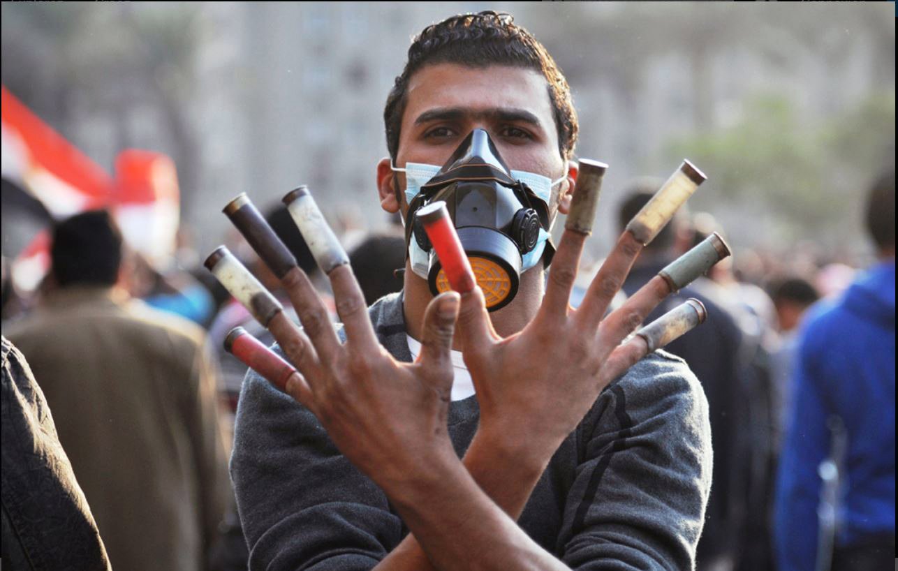 الصورة تظهر شابًا يرتدي كمامة واقية بينما يرفع يديه للتعبير عن موقفه. تظهر أصابعه تحمل أنابيب ملونة، مما قد يشير إلى استخدامه لأدوات معينة في سياق احتجاج أو تجمع شعبي. الخلفية تبدو غير واضحة ولكنها تشير إلى وجود حشد من الناس، مما يدل على أن الصورة قد التقطت في مكان يشهد تظاهرات أو فعاليات سياسية. تعبر الصورة عن مشاعر قوية من المقاومة والتحدي.
