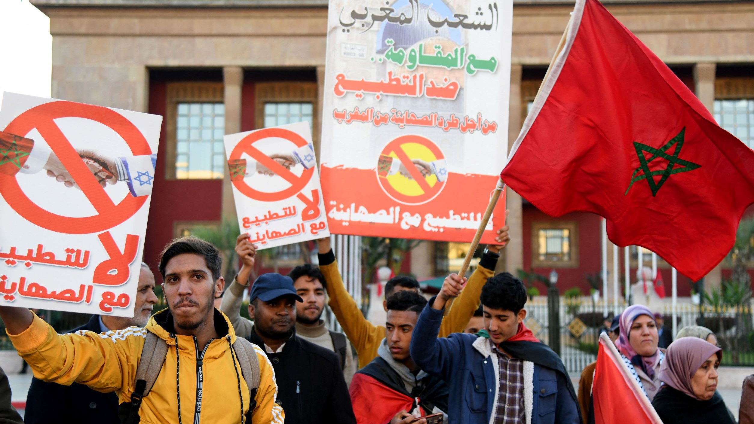 L'image montre un groupe de manifestants tenant des pancartes. Les affiches portent des messages visant à s'opposer à la normalisation des relations avec Israël, avec des symboles qui suggèrent un rejet de cette initiative. Les participants, d'âges divers, expriment leur désaccord à travers des slogans et des gestes. Certains portent des vêtements d'hiver, et l'ambiance semble être celle d'une manifestation pacifique. En arrière-plan, on aperçoit un bâtiment public, probablement lié au gouvernement.