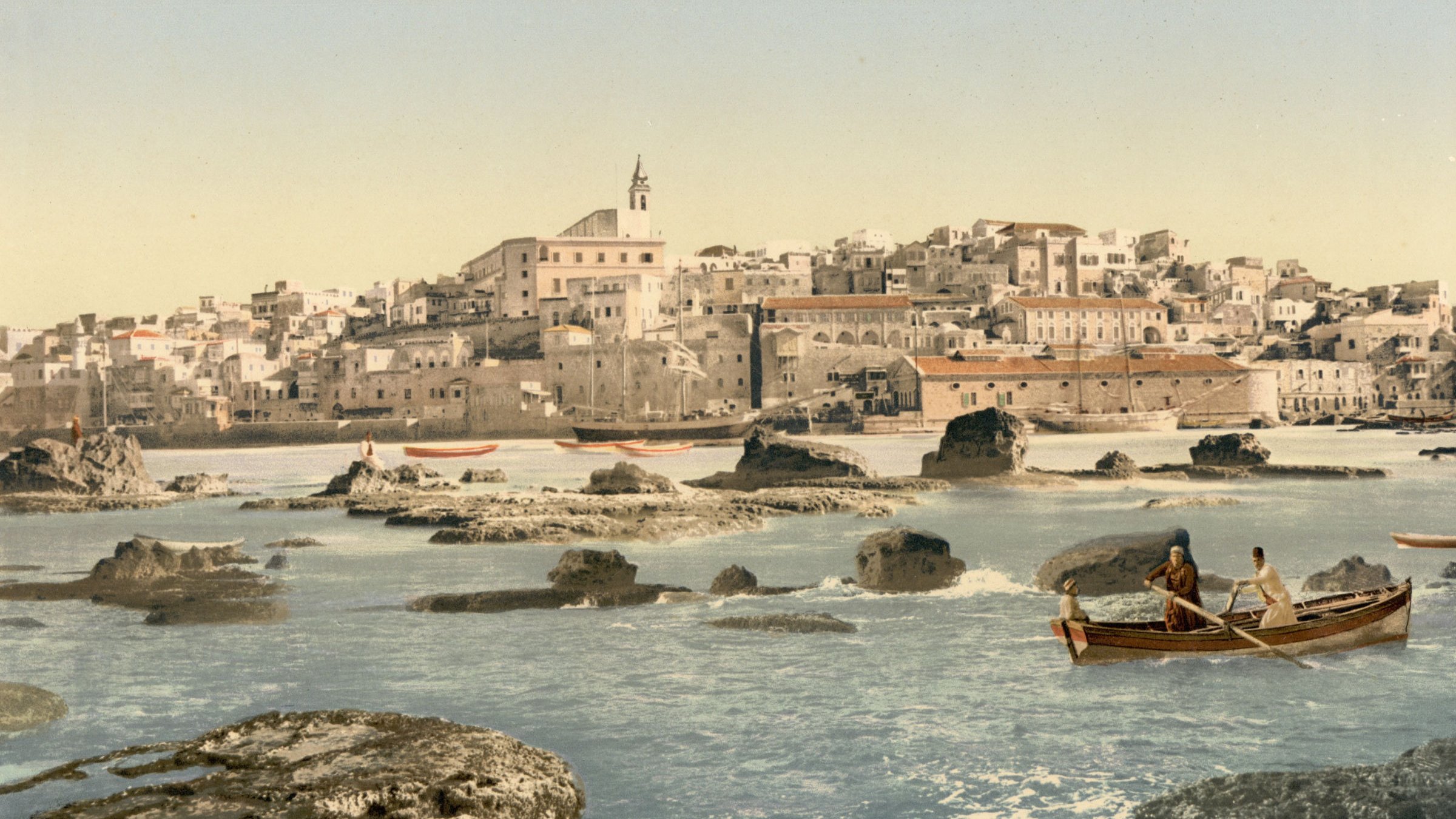 L'image représente un paysage côtier avec une ville en arrière-plan. On peut voir des maisons blanches construisant un front de mer, accompagnées d'une église au sommet d'une colline. Au premier plan, deux personnes naviguent dans un petit bateau sur une eau calme, entouré de rochers. Le ciel est dégagé, suggérant une journée ensoleillée, et la scène évoque une atmosphère paisible et pittoresque. Les couleurs sont vives, mettant en valeur la beauté naturelle de l'endroit.