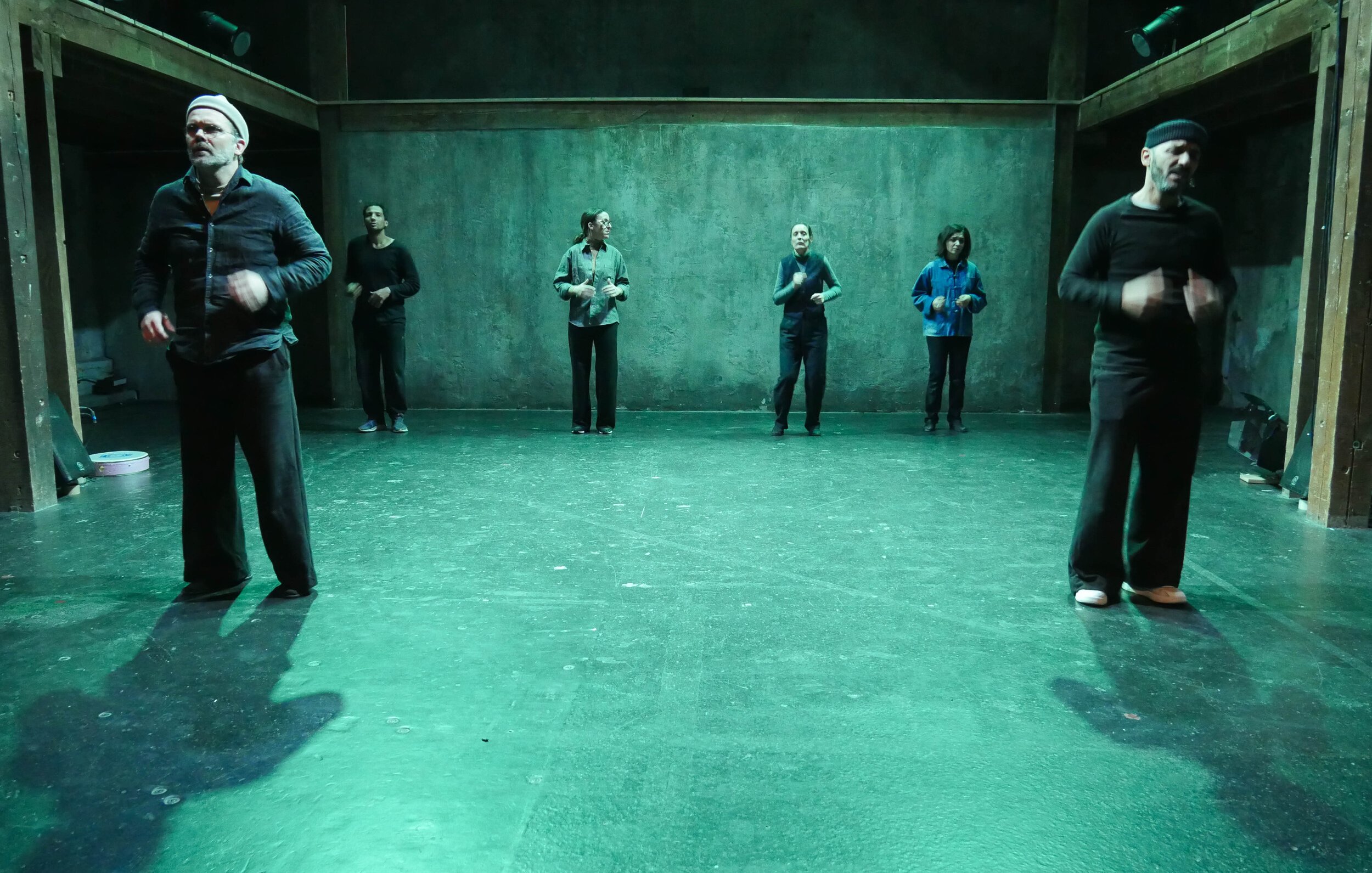 L'image montre un groupe de six personnes sur scène, dans un espace artistique éclairé d'une lumière verte. Elles semblent engagées dans une performance ou une répétition. Les membres du groupe portent des vêtements de différents styles, et se déplacent à divers endroits de la scène. L'arrière-plan est dépouillé, avec des murs en béton, ce qui crée une ambiance minimaliste et contemporaine.