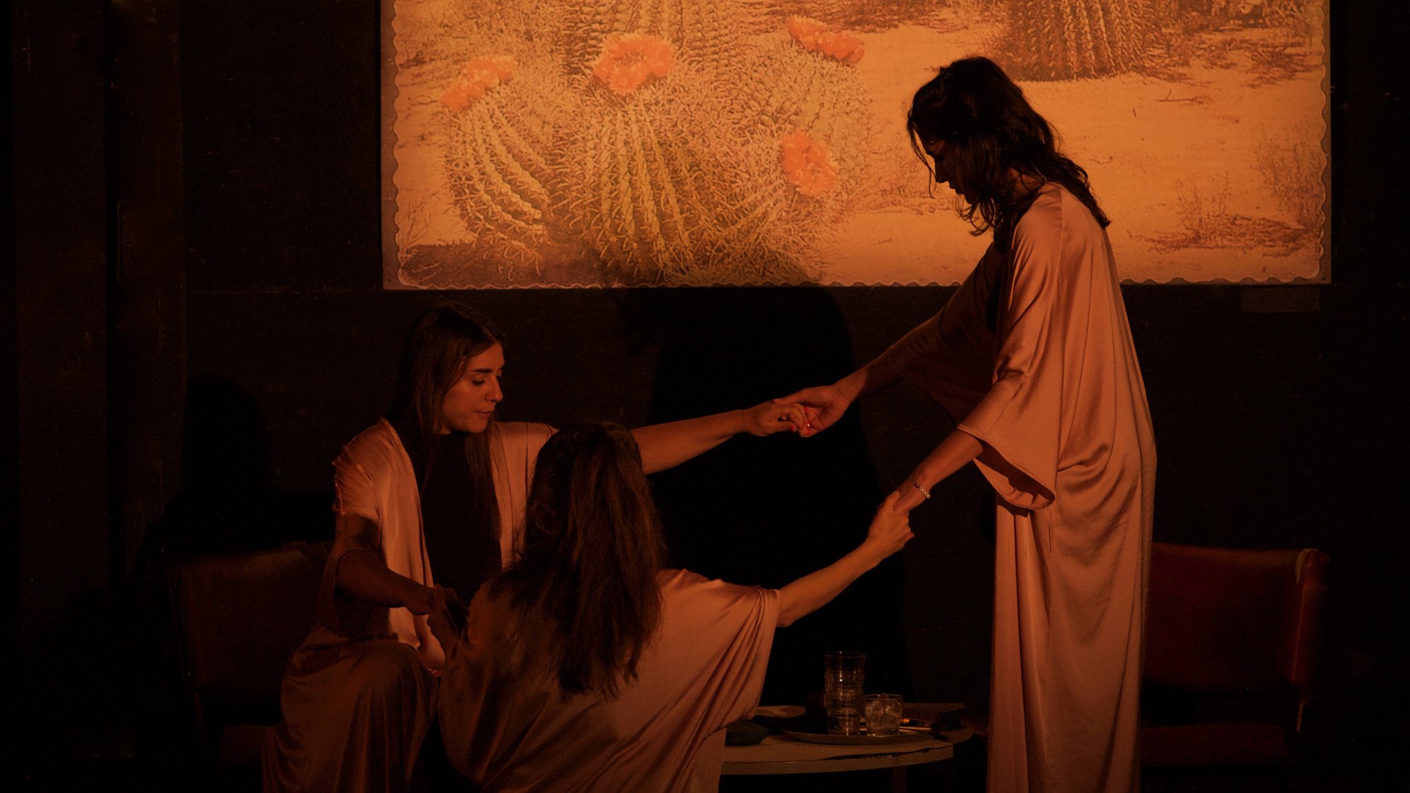 L'image présente une scène théâtrale ou artistique, où trois femmes en vêtements fluides de couleur beige se tiennent autour d'une table basse. Elles semblent engagées dans une interaction attentive, tenant les mains les unes des autres. La lumière est tamisée, créant une atmosphère intime et mystérieuse. En arrière-plan, il y a un tableau représentant des cactus dans un paysage désertique, ajoutant une touche de nature à la scène. Les expressions des femmes ne sont pas clairement visibles, contribuant à l'ambiance intrigante de l'image.