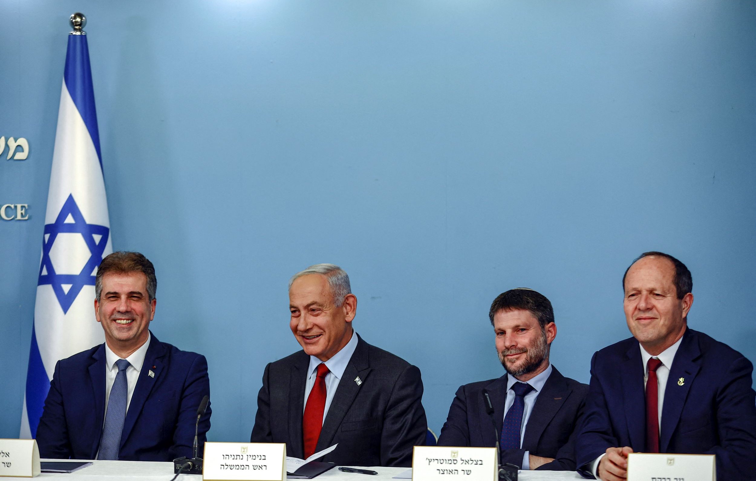 The image shows a group of four men seated at a table in front of a blue backdrop with the Israeli flag. They appear to be engaged in a discussion or announcement. The man in the center, likely the most prominent figure, is smiling. Each individual has a nameplate in front of them, indicating their identities or titles. The overall atmosphere seems formal, suggesting a political or governmental context.