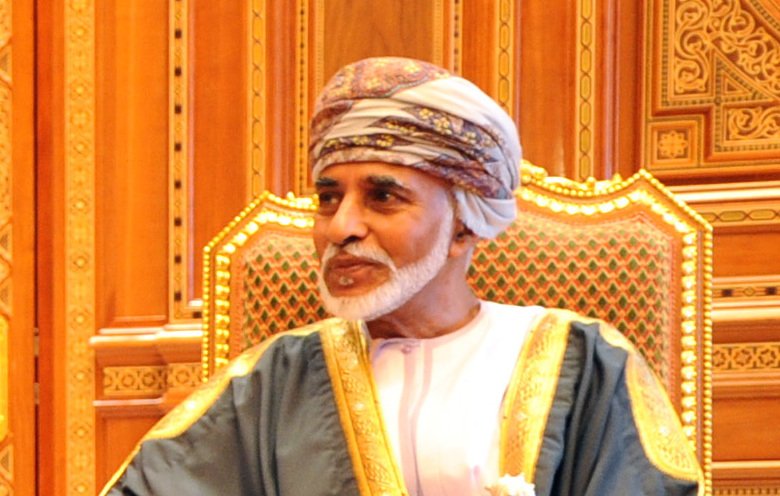 The image features a man seated in a richly decorated chair, wearing traditional attire. He is dressed in a white dishdasha, complemented by a decorative cape, and a turban, which is traditional in some Middle Eastern cultures. The background consists of intricate woodwork and ornate design elements, suggesting an opulent setting. The man's expression appears calm and thoughtful.