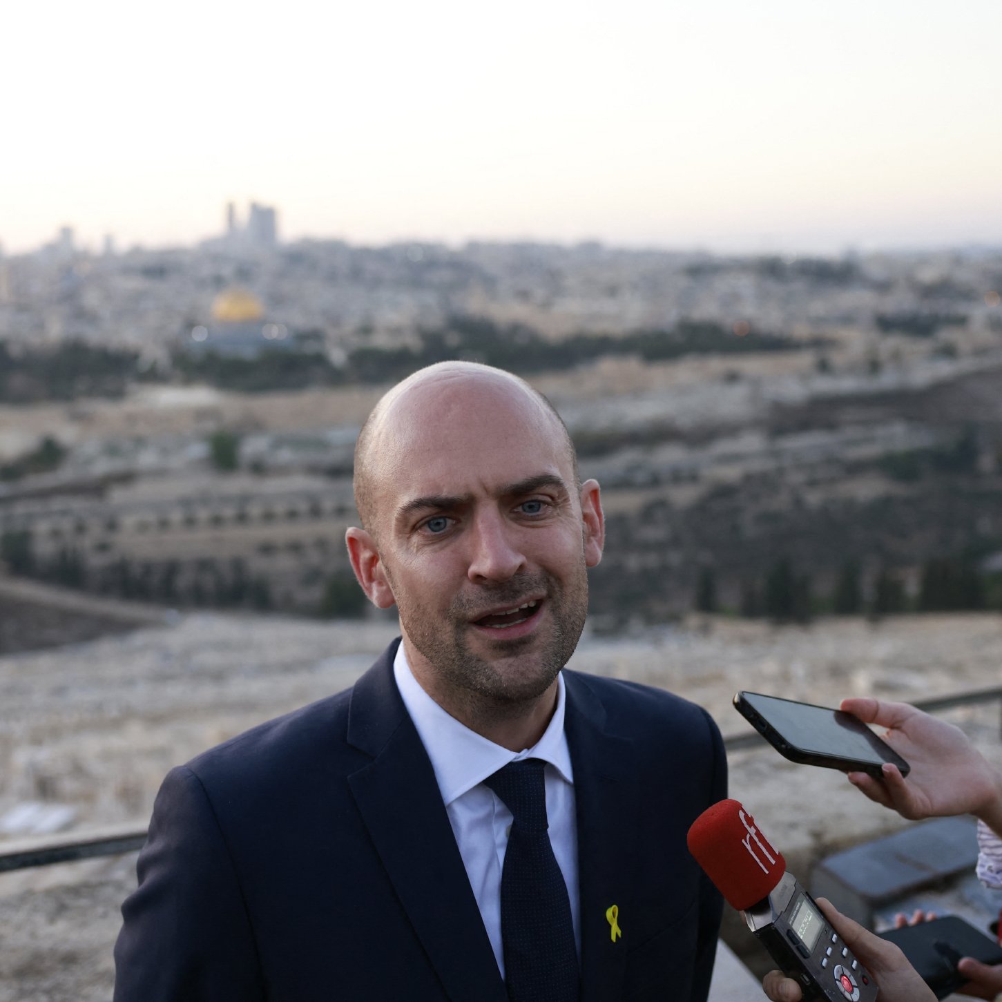 L'image montre un homme en costume, probablement un porte-parole ou un responsable politique, s'exprimant devant un groupe de journalistes. Il se tient à l'extérieur avec une vue panoramique de la ville en arrière-plan, qui semble être Jérusalem, avec des bâtiments et un paysage urbain. Le ciel est clair, et la lumière du crépuscule donne une atmosphère calme à la scène. Plusieurs microphones et téléphones montrent l'intérêt médiatique pour ses déclarations.