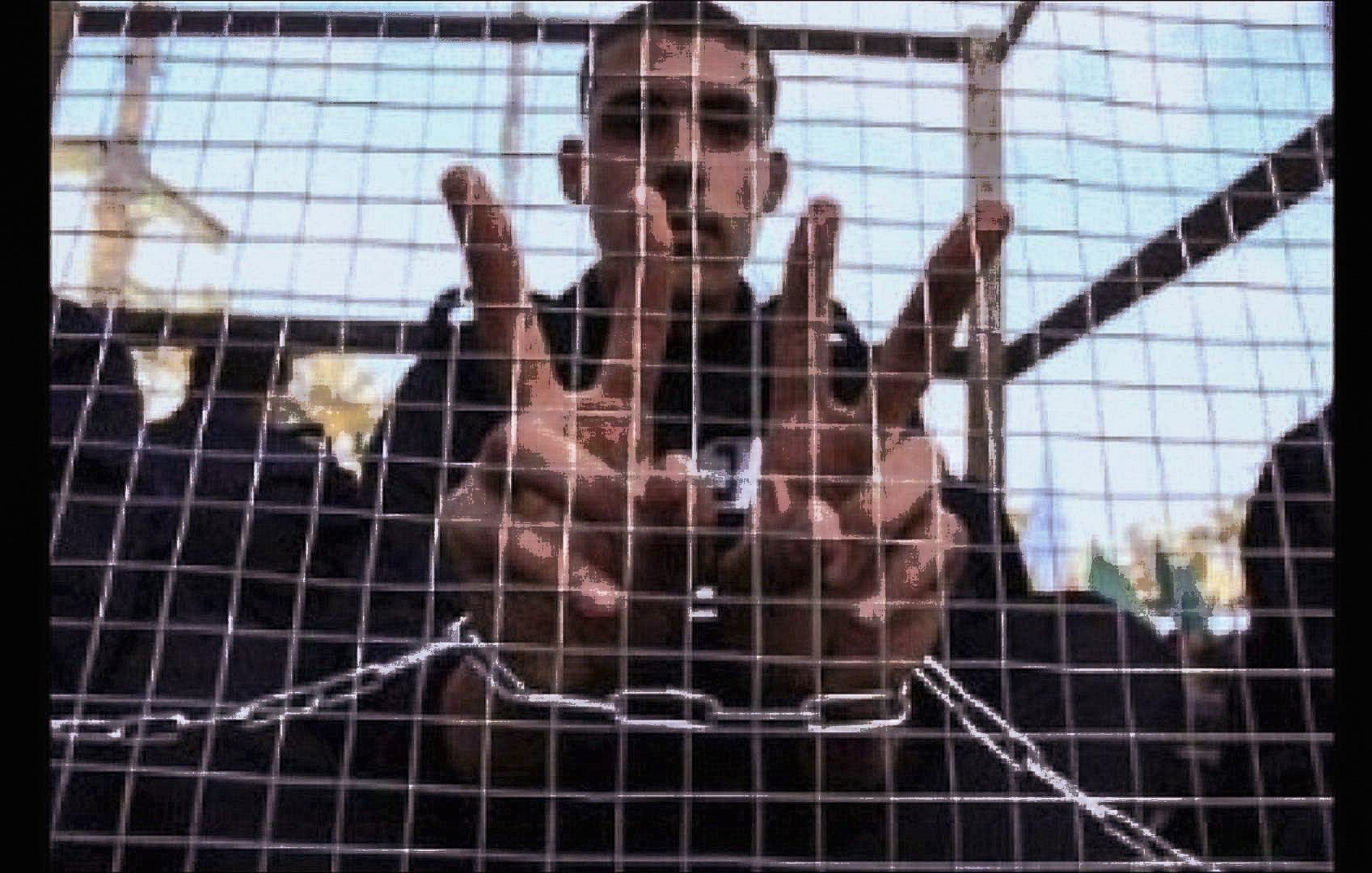 L'image montre une personne derrière une grille ou une structure en métal. Cette personne utilise ses mains pour former un symbole ou un chiffre, probablement en rapport avec un message ou un mouvement spécifique. L'ambiance de l'image semble évoquer une situation de confinement ou de protestation. L'arrière-plan est flou, ce qui met l'accent sur les mains et le geste.