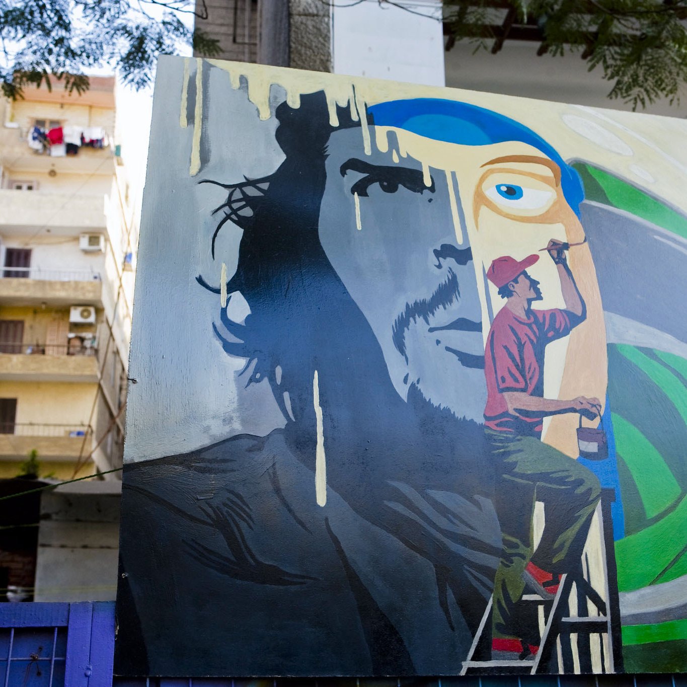 تظهر في الصورة لوحة فنية كبيرة على حائط، تمثل وجه رجل بشكل جزئي، حيث يتواجد شخص آخر على سُلّم يرسم تفاصيل إضافية. الخلفية تحتوي على ألوان زاهية، مع تجسيد ذوات رموز فنية. كما يُمكن رؤية بعض الشقق السكنية في الخلف بالإضافة إلى الملابس المعلقة على الشرفات. تعكس الصورة أجواء حضرية وفنية.
