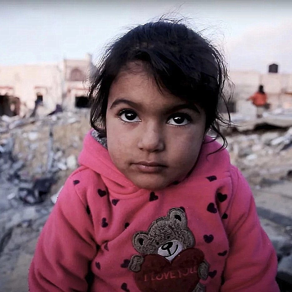 L'image montre une petite fille avec un regard expressif, vêtue d'un sweat à capuche rose orné d'un ours. Elle se trouve dans un environnement dévasté, entourée de décombres et de bâtiments en ruines. En arrière-plan, on aperçoit d'autres enfants. L'atmosphère semble chargée d'une grande tristesse et d'une vulnérabilité palpable.
