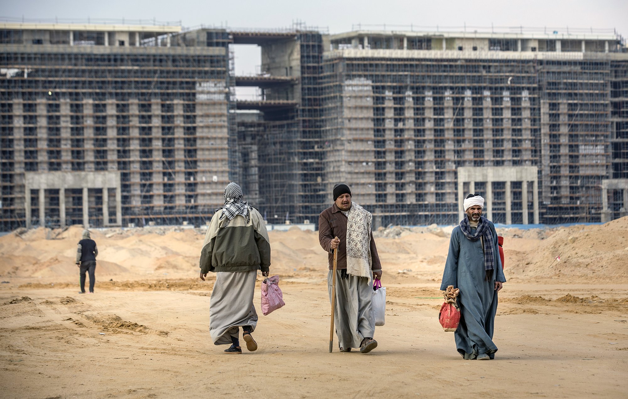 L'image montre un paysage de construction, avec plusieurs bâtiments inachevés en arrière-plan. Au premier plan, on voit quatre personnes marchant sur un sol de terre. Elles portent des vêtements traditionnels et semblent transporter des sacs. L'environnement est désertique, avec des matériaux de construction visibles autour. Le ciel est nuageux, contribuant à l'atmosphère de travail en cours.