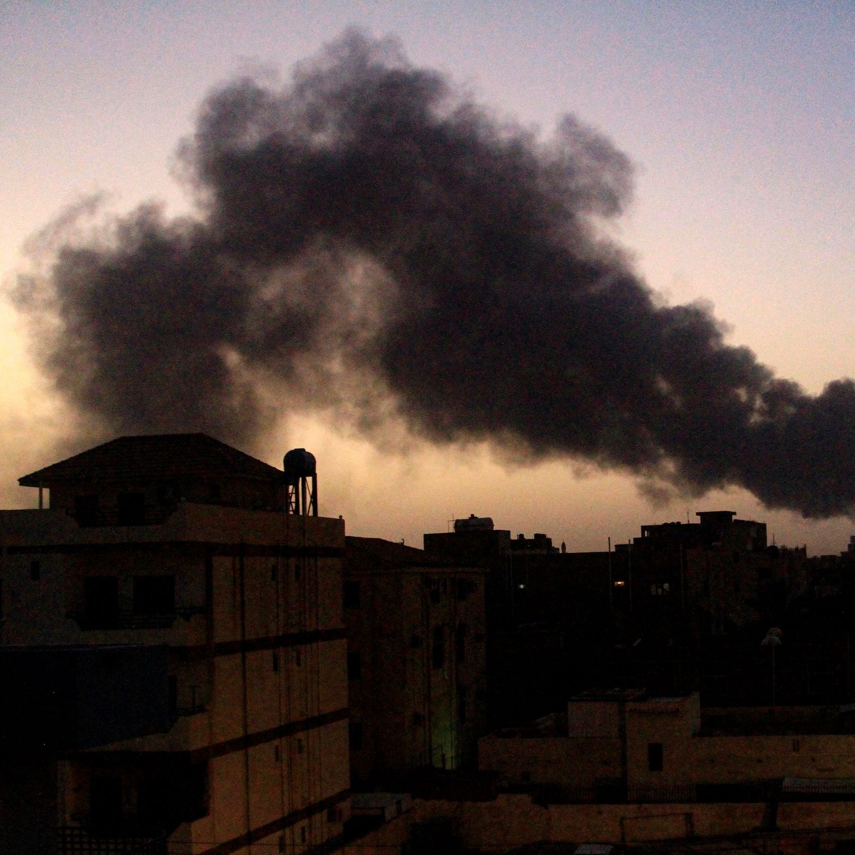 La imagen muestra un paisaje urbano al atardecer, con un cielo que presenta tonos oscuros. En el fondo, se puede ver una densa columna de humo negro que se eleva, sugiriendo la presencia de un incendio o alguna actividad industrial. A la izquierda, hay edificios que se recortan contra el cielo, y a la derecha, unas palmeras iluminadas por luces artificiales. La atmósfera parece tensa debido al humo y el entorno oscuro.