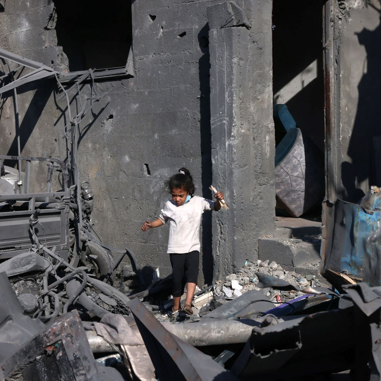 تظهر الصورة طفلاً صغيراً يرتدي الملابس البيضاء وهو يسير بين أنقاض مبنى مدمّر. الجدران حوله متهدمة ومغطاة بالرماد، مما يدل على الدمار الذي حل بالمكان. في الخلفية، يمكن رؤية شخص بالغ يقف بالقرب من الباب، يبدو وكأنه يتفحص وضع المكان. الأجواء تعكس حالة من الحزن والفوضى نتيجة الكارثة.