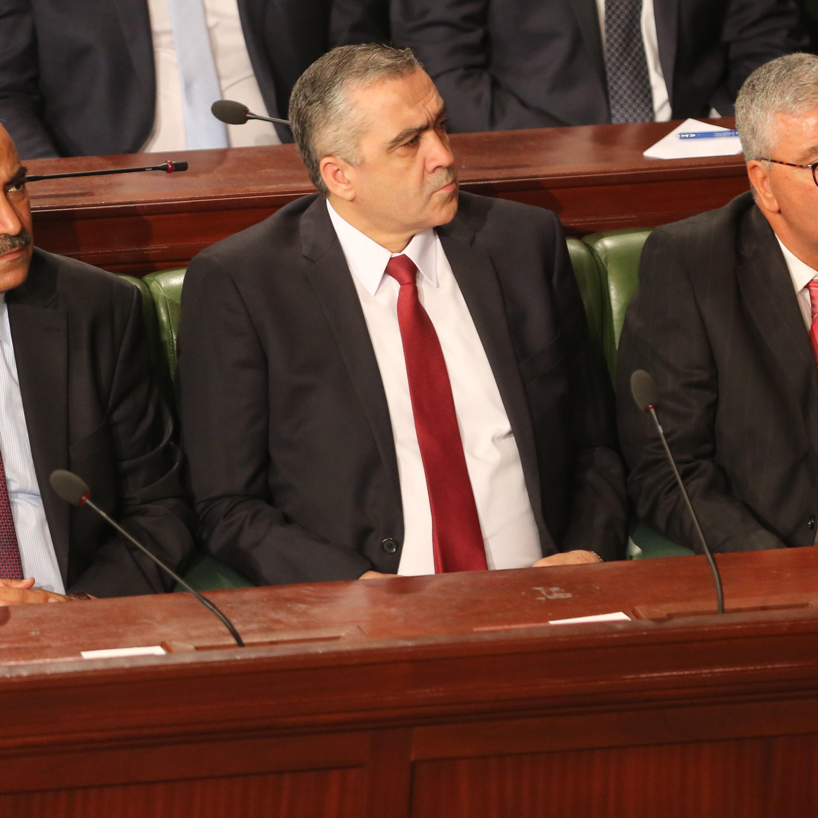 The image shows three men sitting side by side in a formal setting, likely within a legislative or governmental assembly. They are dressed in business suits, with two wearing ties—one in red and the other in a lighter color. The background suggests a gathering or meeting, with a group of people seated behind them. The scene conveys a sense of seriousness and official duty.