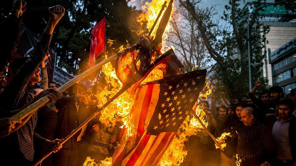 L'image montre une scène de protestation où des gens brûlent un drapeau américain. Les participants, visiblement passionnés, lèvent les bras et brandissent des bâtons. L'atmosphère est intense, avec des flammes qui s'élèvent autour du drapeau, tandis que des arbres et des bâtiments en arrière-plan ajoutent à la dynamique de la situation. Cette action symbolise souvent une forte opposition ou un message politique.