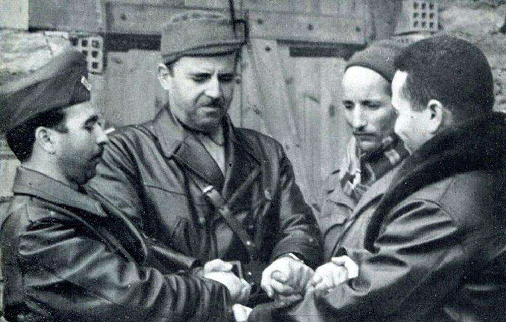 L'image montre quatre hommes serrant les mains les uns des autres dans une posture de camaraderie ou de négociation. Ils portent des tenues militaires ou de combat, et l'environnement semble être extérieur, avec des murs en pierre en arrière-plan. L'expression de leurs visages peut suggérer la détermination ou la concentration, et l'interaction semble exprimer un moment significatif.