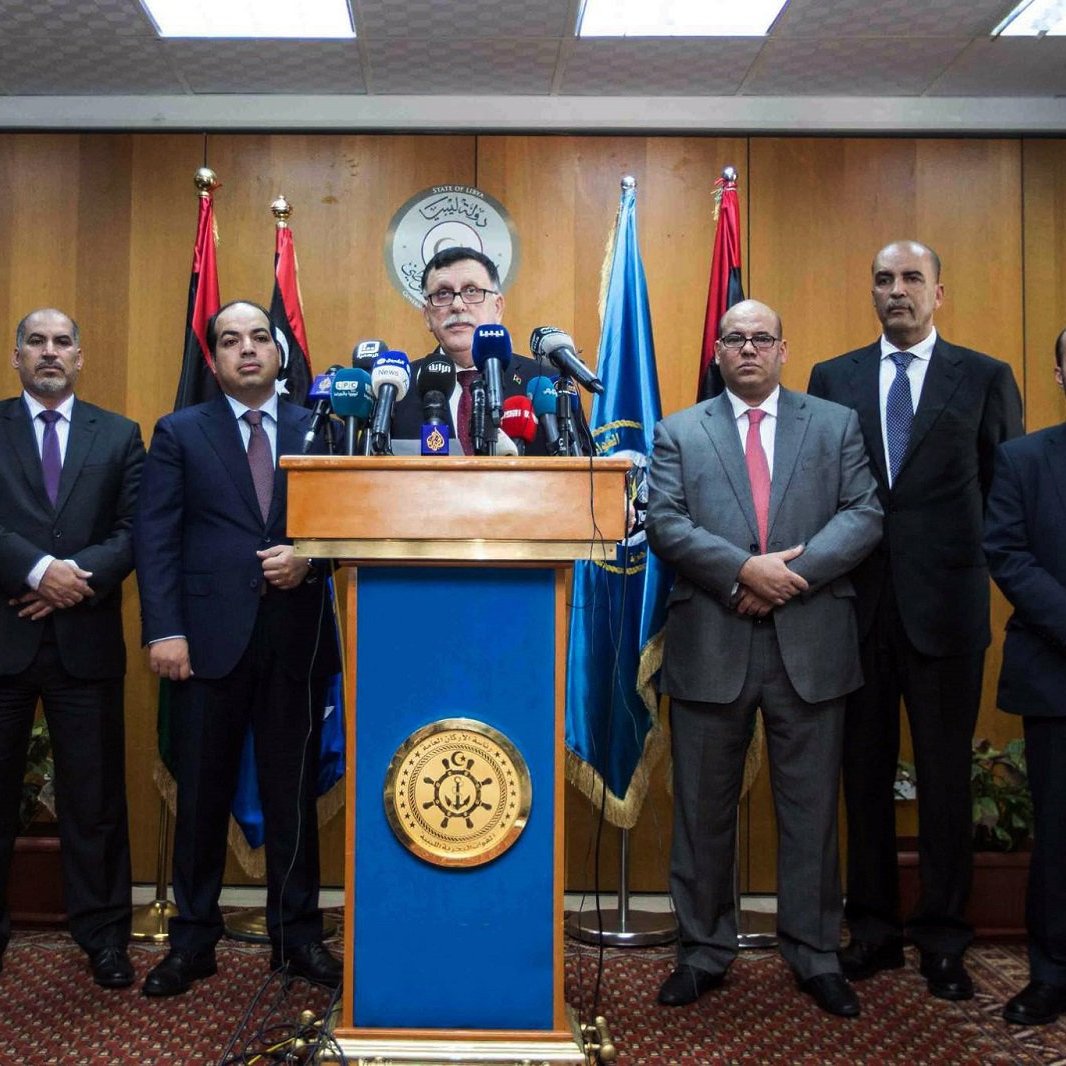 The image shows a group of eight men standing in a formal setting, likely a press conference or official announcement. There is a podium at the center with a seal or emblem, and the speaker appears to be addressing the audience. The backdrop features flags, suggesting a governmental or institutional context. The men are dressed in suits and ties, conveying a professional atmosphere.