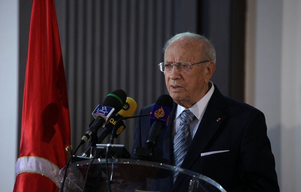 L'image montre un homme âgé qui se tient derrière un pupitre avec des micros. Il porte un costume sombre et une cravate, et semble s'adresser à un public. En arrière-plan, on peut voir un drapeau rouge, ce qui pourrait indiquer un contexte officiel, possiblement politique. L'atmosphère semble sérieuse et formelle.