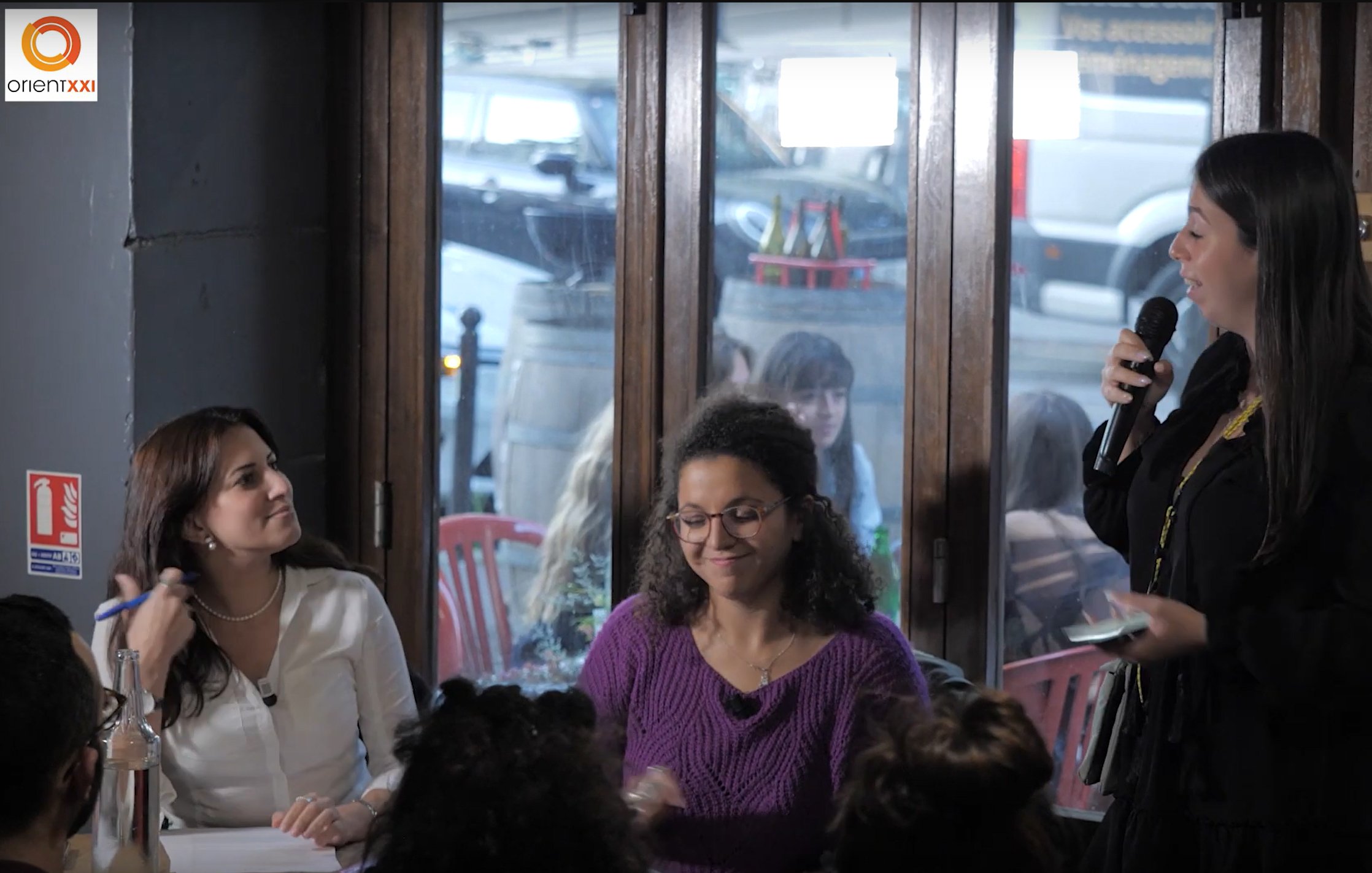 L'image montre une scène intérieure où plusieurs personnes sont assises à des tables. Une femme se tient debout au premier plan, tenant un microphone, probablement en train de parler ou de présenter quelque chose. Elle porte des vêtements noirs et semble engagée dans une conversation dynamique. À sa gauche, une autre femme sourit, tandis qu'à sa droite, une femme avec des lunettes apporte une expression de concentration. À l'arrière-plan, on peut voir des clients assis, créant une atmosphère animée. Les grandes fenêtres laissent entrer la lumière et montrent une vue partielle de l'extérieur.