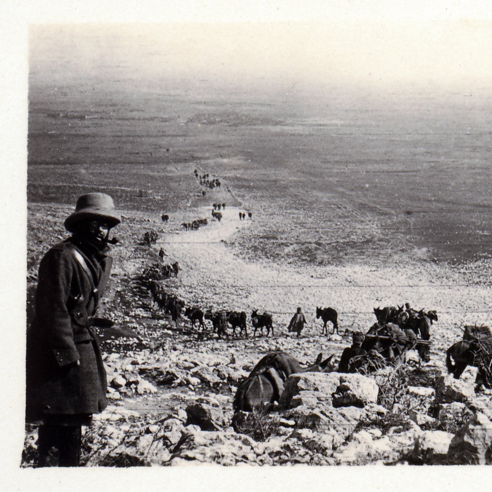 در این تصویر، مردی با کلاه و پوشاک سنتی در حال ایستادن در کنار یک شیب سنگی دیده می‌شود. در پس‌زمینه، گروهی از انسان‌ها و حیوانات (احتمالاً خرها یا اسب‌ها) در حال حرکت در یک مسیر سنگی به سمت پایین هستند. مناظر اطراف خشک و کوهستانی به نظر می‌رسد و تصویر حالتی قدیمی و سیاه و سفید دارد که به آن احساس تاریخ را القا می‌کند. فضای تصویر و نوع پوشش افراد نشان‌دهنده یک فعالیت یا سفر است.