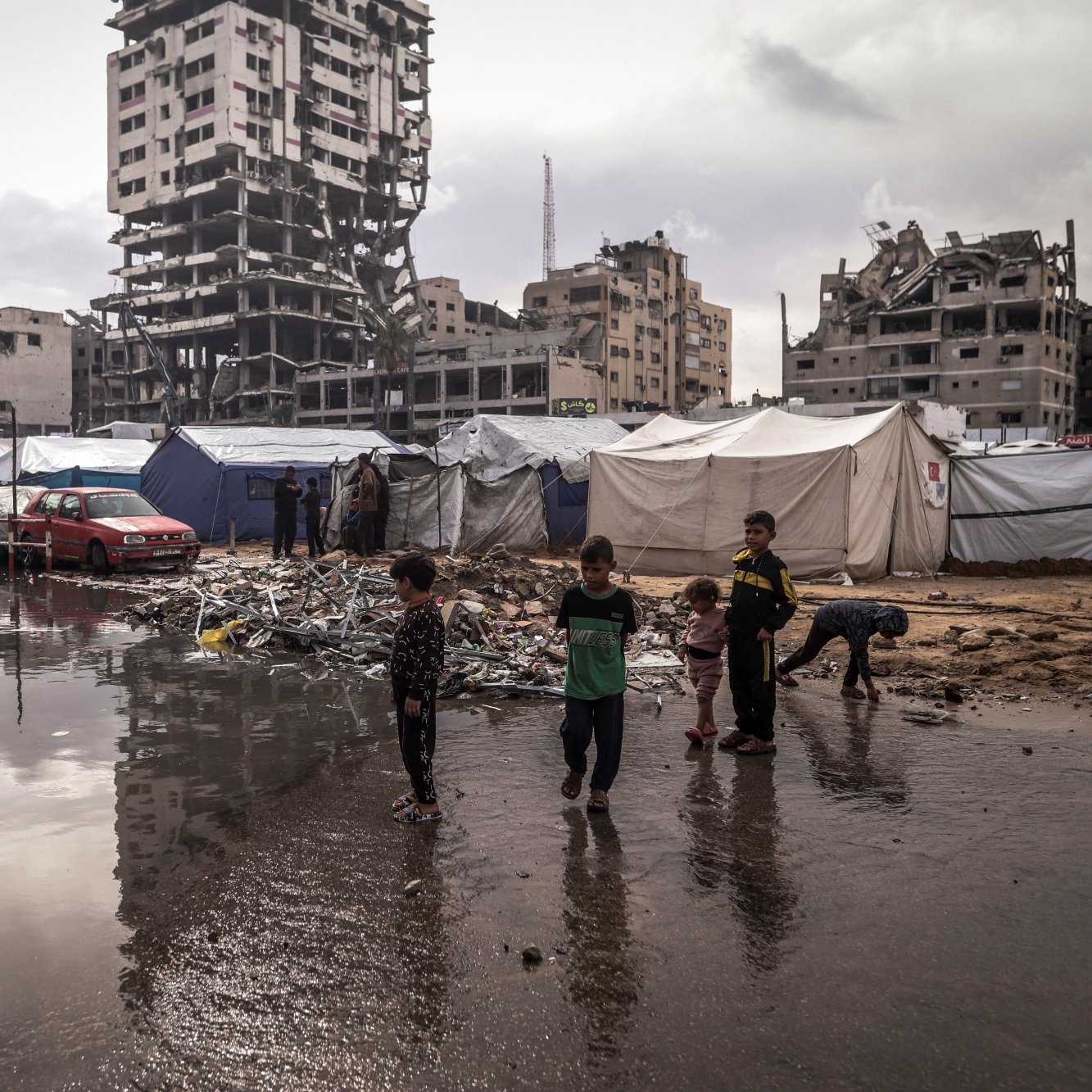Des enfants jouent près de tentes dans un camp, avec un bâtiment en ruines en arrière-plan.