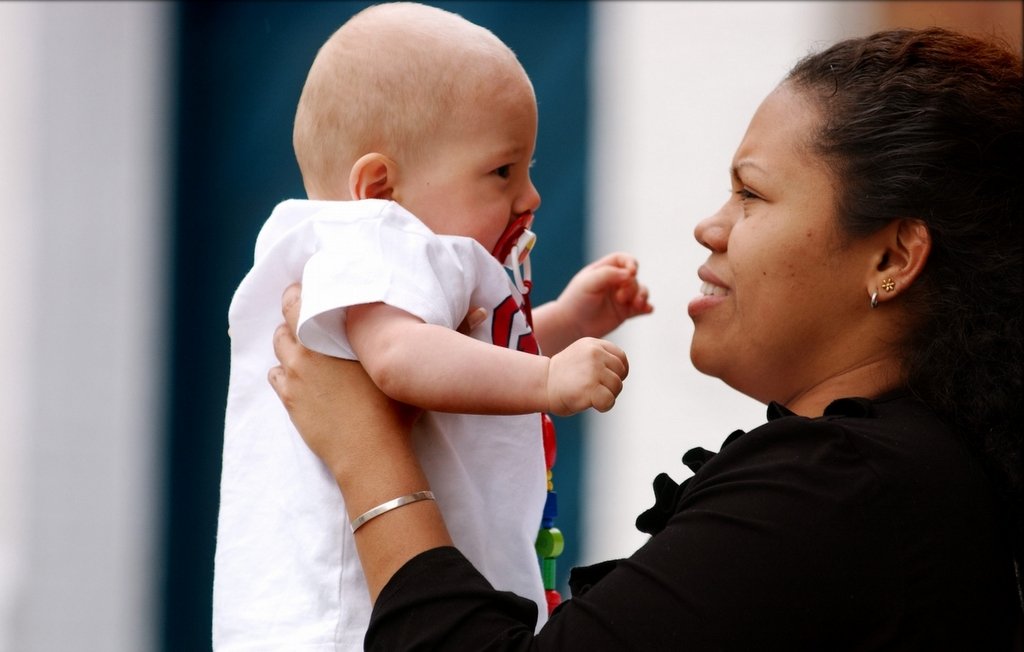 L'image montre une femme tenant un bébé dans ses bras. Le bébé, qui a une tétine, regarde la femme avec curiosité, tandis que celle-ci lui sourit. La femme porte un haut noir et a des boucles d'oreilles. L'arrière-plan est flou et coloré, suggérant un environnement en extérieur. L'atmosphère de la scène est affectueuse et chaleureuse.