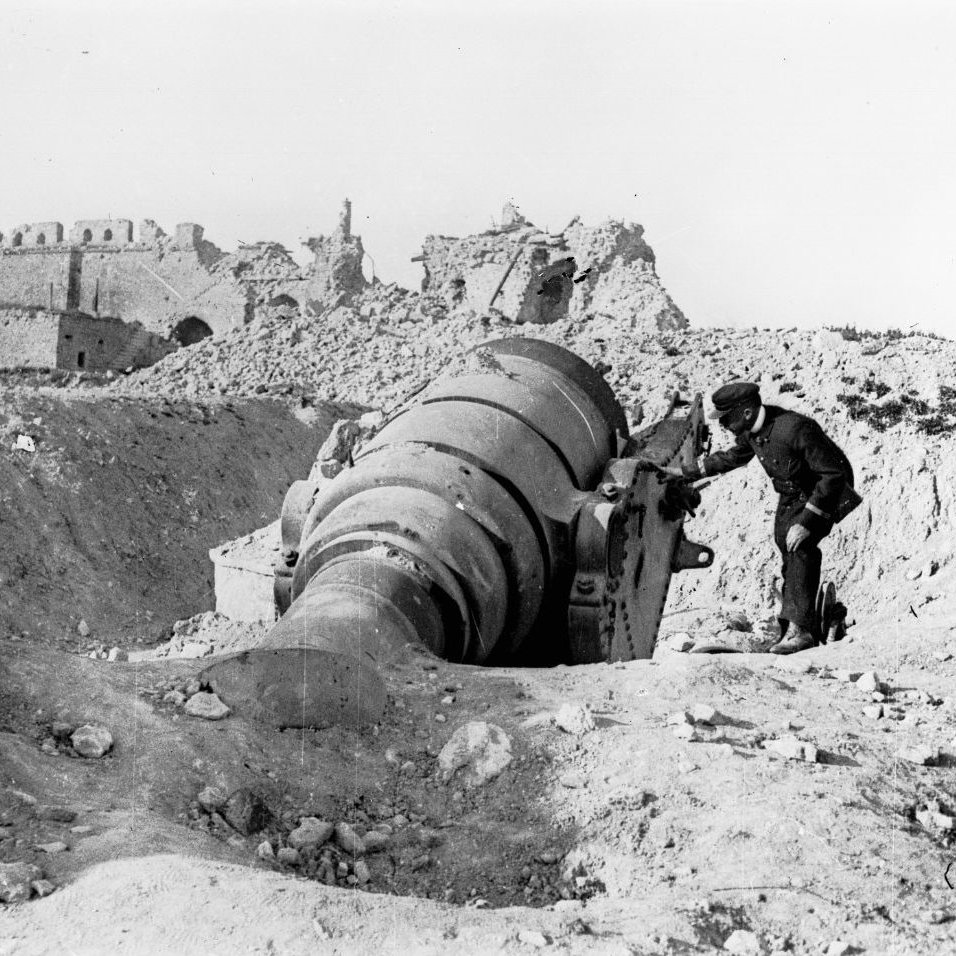 این تصویر به نظر می‌رسد نمایی تاریخی از یک مکان جنگی باشد. در تصویر، یک مرد به یک توپ بزرگ که به سمت زمین خوابیده است، نزدیک می‌شود. پس‌زمینه شامل ویرانه‌هایی از ساختمان‌ها یا قلعه‌های قدیمی است که گویای تخریب ناشی از جنگ یا نبردی است. همچنین، زمین اطراف آن سنگ‌ریزه‌ها و خاکی دارد که نشان‌دهنده شرایط ناهموار و دشوار می‌باشد. به طور کلی، این تصویر حس غم و خرابی را منتقل می‌کند و یادآور هزینه‌های جنگ است.