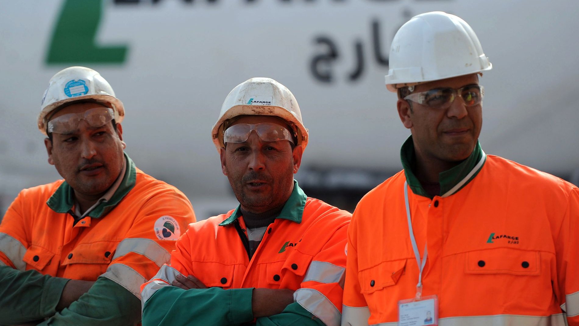 L'image montre trois ouvriers portant des vêtements de sécurité orange et des casques blancs. Ils se tiennent debout avec les bras croisés, affichant une attitude sérieuse. En arrière-plan, on peut voir un camion ou un véhicule avec le logo de Lafarge, une entreprise connue dans le secteur des matériaux de construction. L'environnement semble être un chantier ou une zone industrielle.