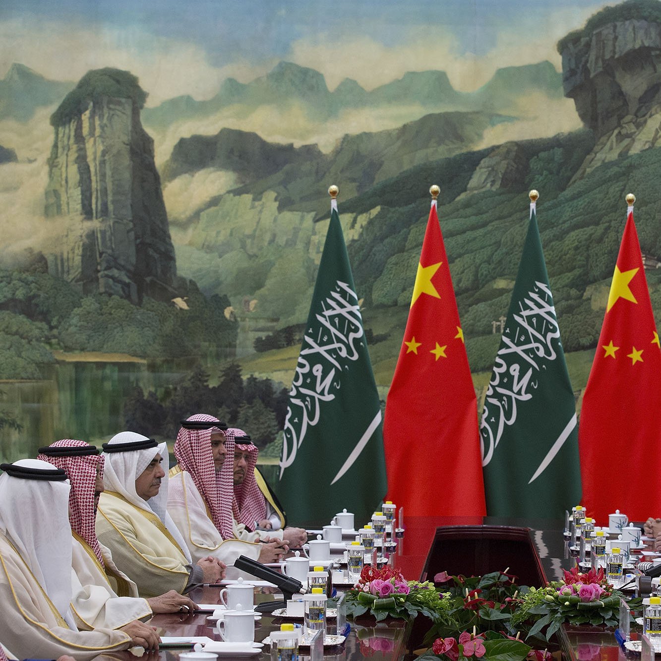 در این تصویر، یک نشست رسمی بین مقامات سعودی و چینی در حال برگزاری است. دو طرف میز مذاکره نشسته‌اند و پرچم‌های عربستان سعودی و چین به وضوح در پس‌زمینه دیده می‌شود. فضای جلسه به گونه‌ای طراحی شده که یک پس‌زمینه طبیعی با کوه‌ها و درختان وجود دارد. هر طرف میز با تعداد زیادی فنجان و ظروف غذا چیده شده است و به نظر می‌رسد که محتوای بحث و تبادل نظر جدی و مهم باشد.
