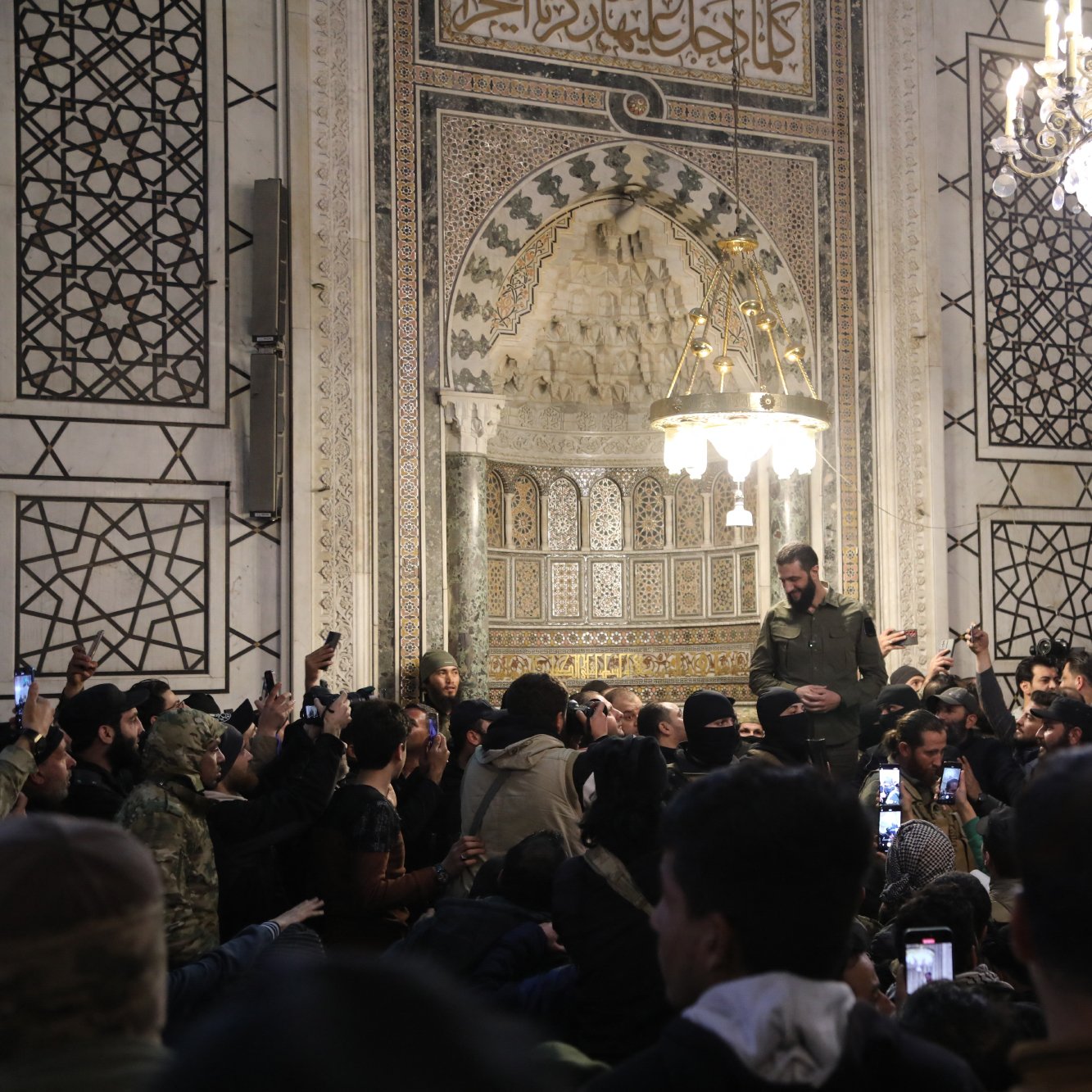 The image depicts a gathering inside a beautifully decorated building with intricate architectural details. The hall features ornate tiles and a grand chandelier hanging from the ceiling. A crowd of people is visible, some holding up their smartphones to capture the moment. In the center, a man dressed in military-style attire stands on an elevated platform, addressing the audience. The atmosphere seems to be one of engagement and attentiveness, with many individuals focused on the speaker.