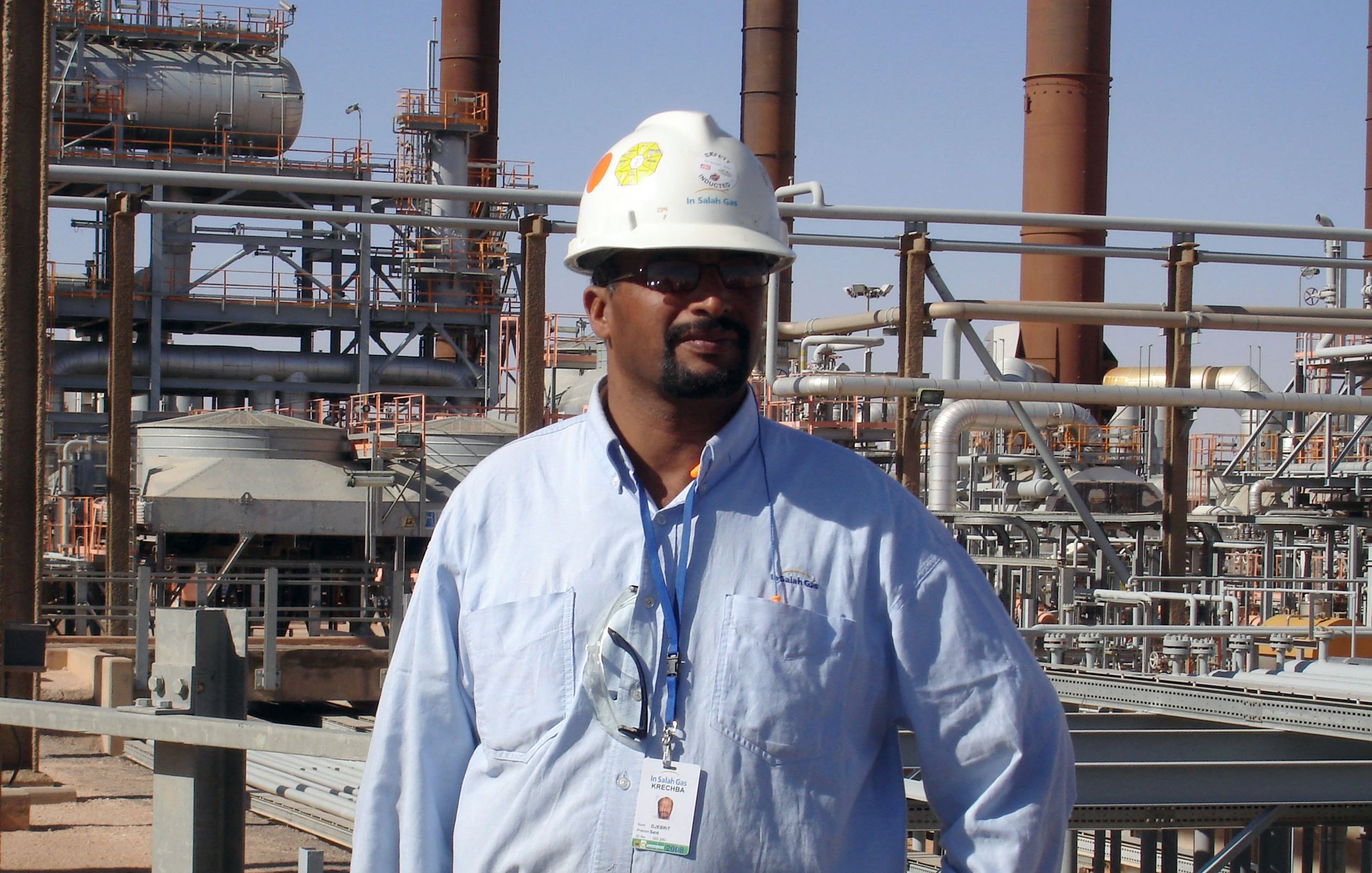 L'image montre un homme portant un casque de sécurité blanc et une chemise bleue, debout dans un environnement industriel. En arrière-plan, on aperçoit des structures métalliques et des installations, probablement liées à une usine ou une raffinerie. L'homme semble professionnel et attentif, avec une carte d'identification suspendue à son cou.