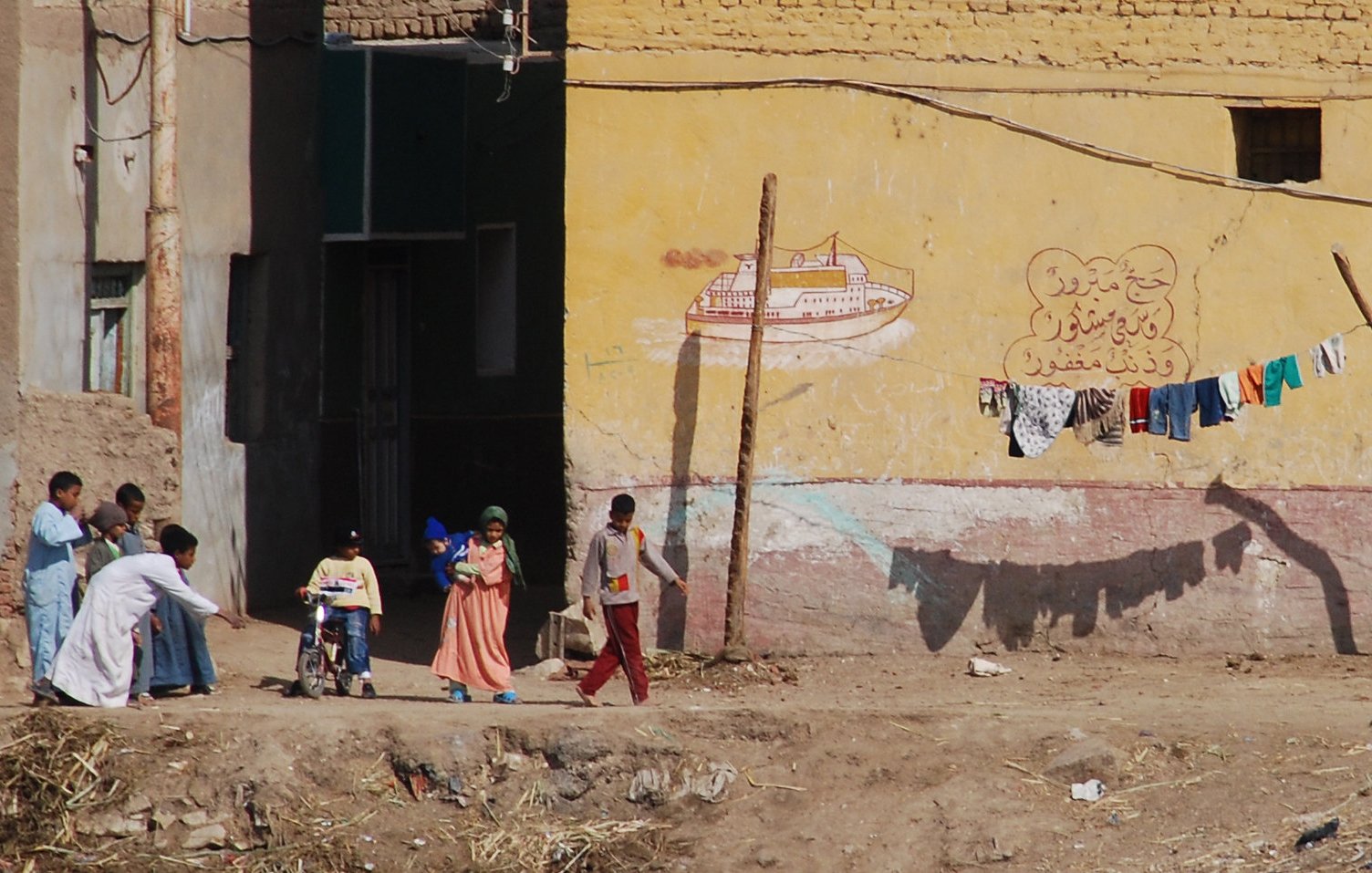 L'image montre une scène de rue dans un environnement rural ou semi-urbain. On peut y voir plusieurs personnes, dont des enfants, qui se déplacent. À l'arrière-plan, il y a des bâtiments avec des murs peints, incluant des motifs ou des inscriptions. Des vêtements sont suspendus à une corde, presque comme un étendage. Le sol est poussiéreux, et l'ensemble de la scène dégage une atmosphère de vie quotidienne dans un cadre modeste.