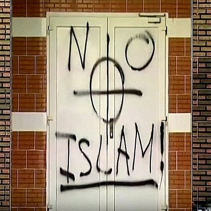 تظهر في الصورة بابًا مغطى بكتابات جدارية، حيث تم رسم عبارة "لا إسلام" مع رمز يظهر قوسًا دوائريًا. الكتابة تبدو باللون الأسود على خلفية بيضاء. الجدران المحيطة بالباب مصنوعة من الطوب بلون بني فاتح. الصورة تعكس مشاعر سلبية تجاه الإسلام، مما يثير جدلاً حول التسامح والقبول.