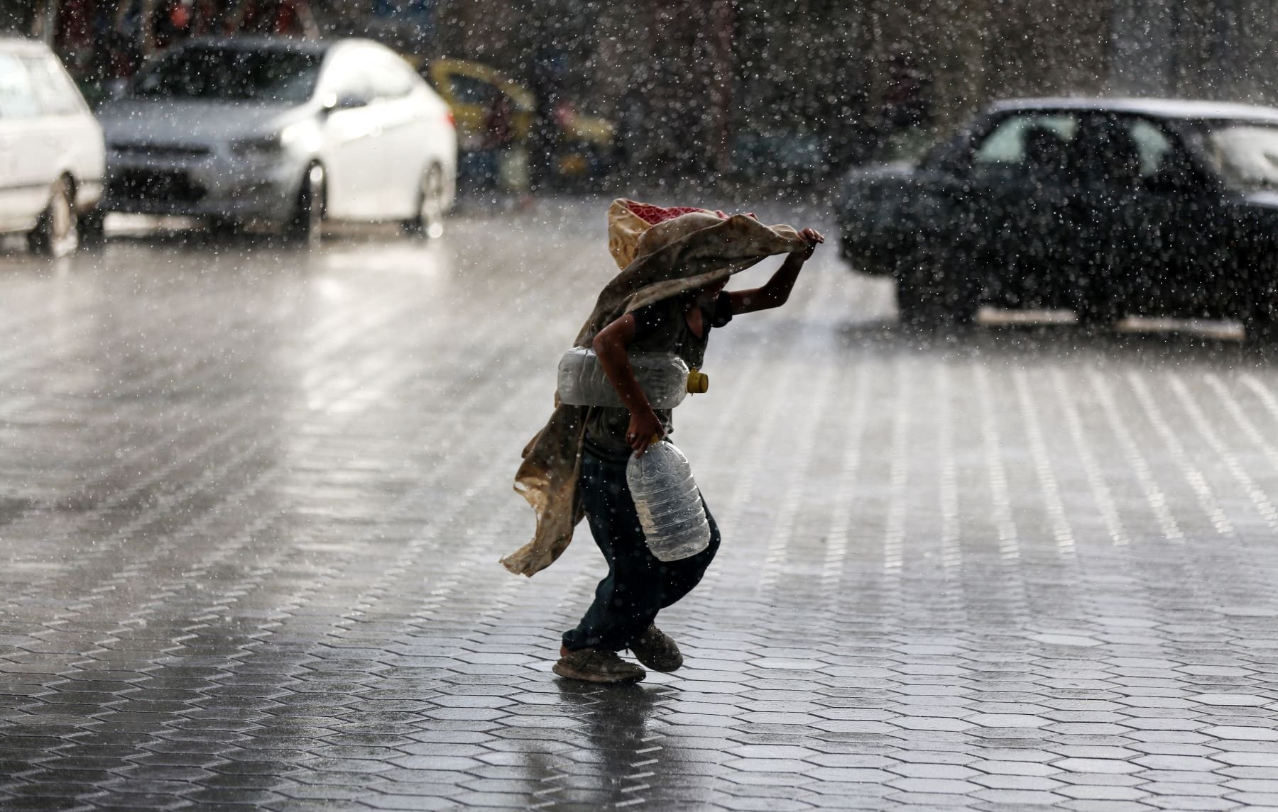 L'image montre un enfant courant sous la pluie intense, portant une écuelle en métal et un sac. Il semble se protéger de la pluie avec un tissu qui couvre sa tête et ses épaules. Le sol est mouillé et réfléchit la lumière, tandis que l'environnement autour de lui est flou en raison des cordes de pluie. Des voitures sont visibles en arrière-plan, accentuant l'atmosphère urbaine de la scène.