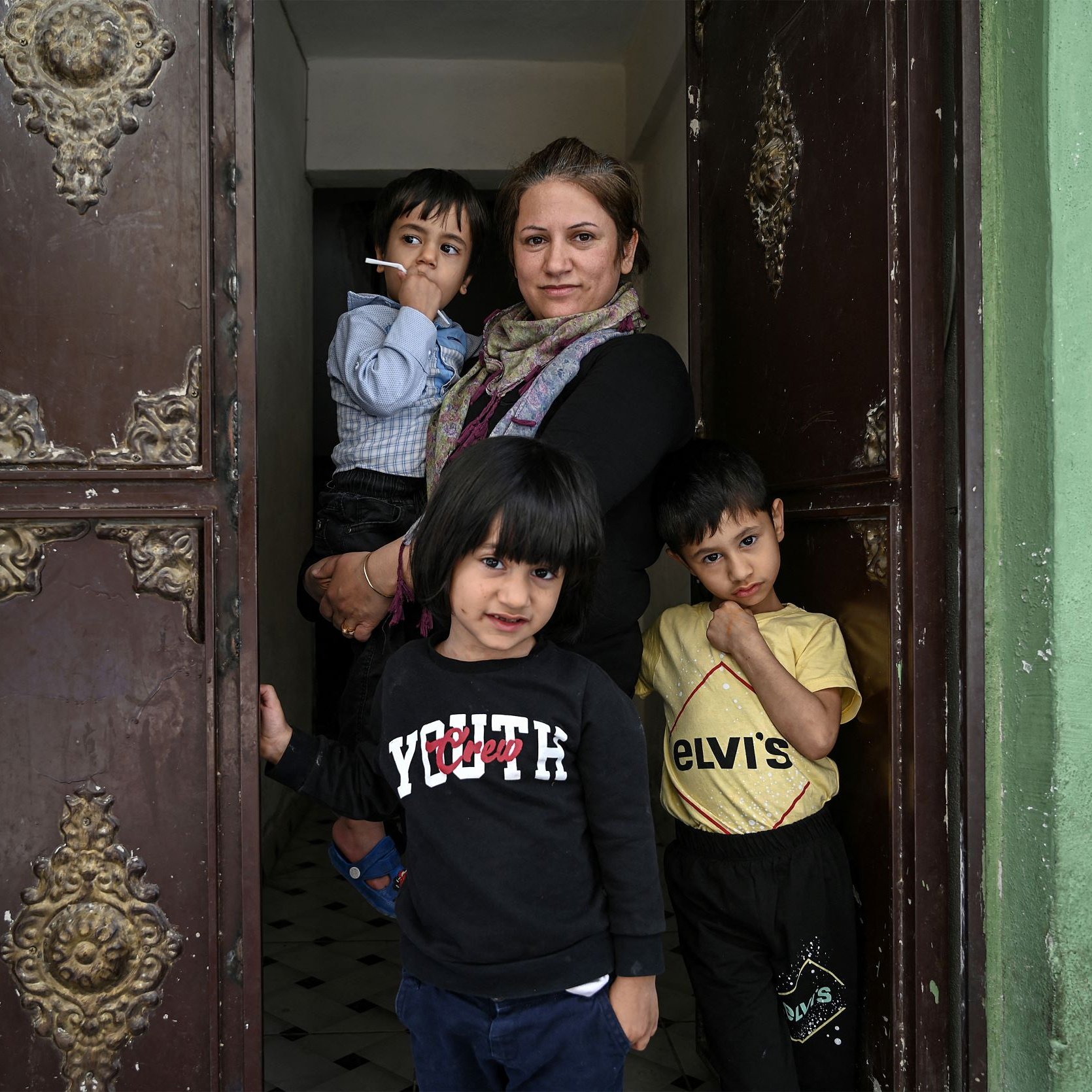 The image depicts a woman standing in a doorway with three young children. The woman appears to be holding a cigarette and has a warm expression. The children are posed around her; two are standing behind her in the doorway, while one child is in front, looking directly at the camera. The surroundings have a vintage feel, with decorative doorwork and green walls. The overall atmosphere reflects a sense of family and community.