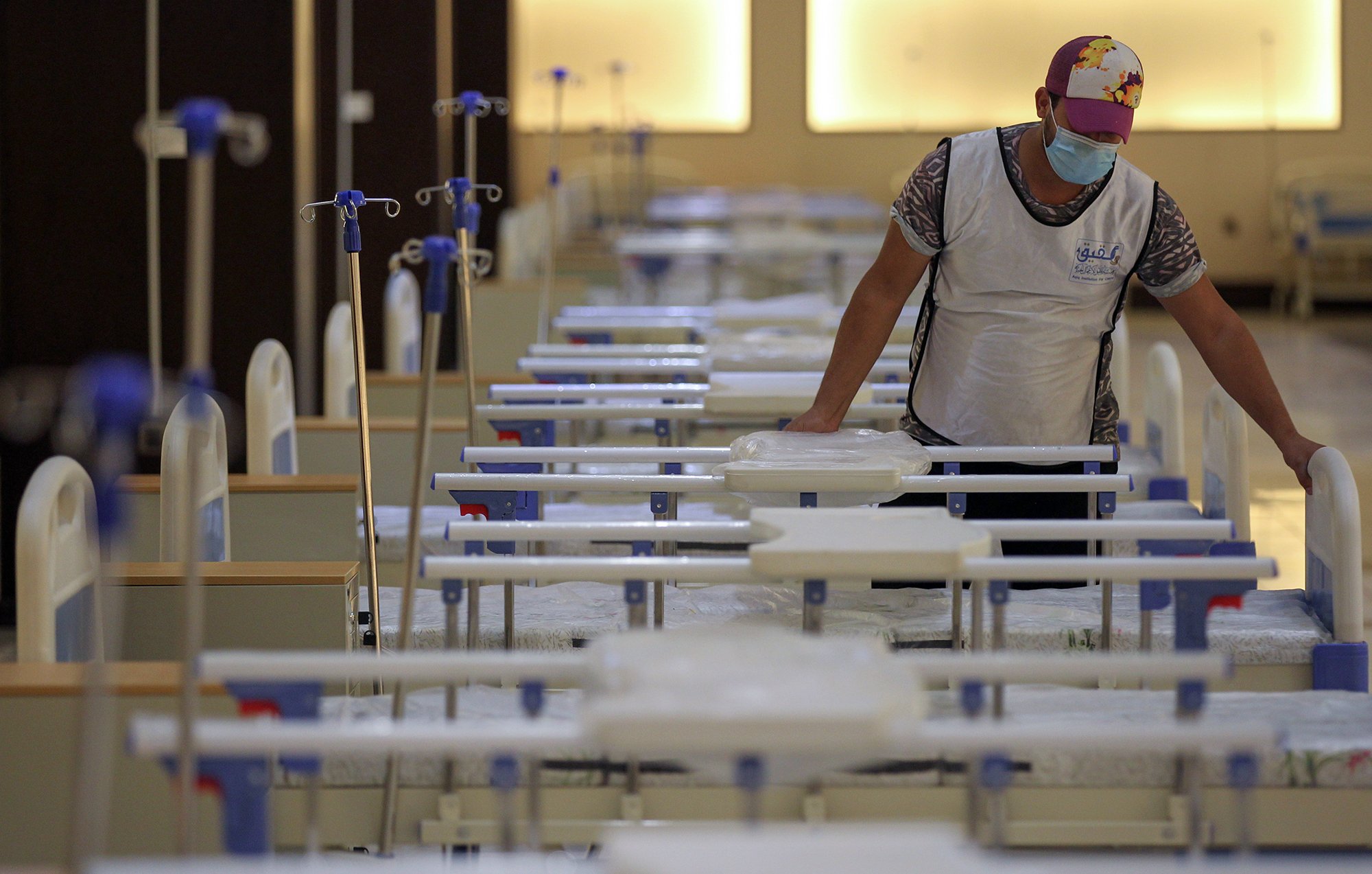 The image shows a person in a mask and a cap standing in a room filled with hospital beds. The beds are arranged in neat rows, and the area appears organized and prepared, possibly for medical purposes. The individual is interacting with the beds, which may indicate they are setting up or maintaining the space. The lighting in the room is warm and soft, contributing to a calm atmosphere.