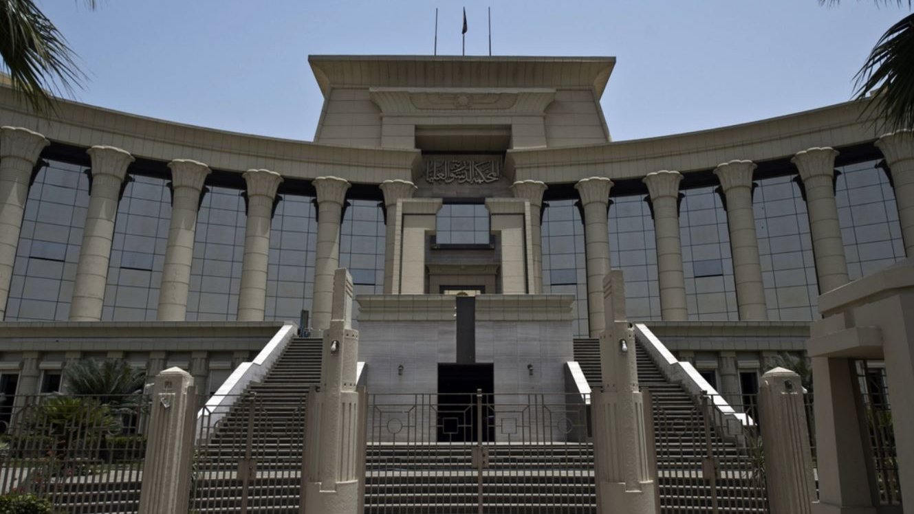 الصورة تظهر مبنى حكومي كبير يتميز بتصميم معماري حديث. يبرز في الصورة مدخل المبنى الذي يحتوي على مجموعة من السلالم والأعمدة الكبيرة. الواجهة تمتاز بالنوافذ الزجاجية العريضة التي تعكس الضوء، وتوجد أعلام مرفوعة فوق السطح. المنطقة المحيطة بالمبنى تحتوي على أشجار ونباتات تعطي جواً من الخضرة. يبدو الطقس مشمساً وواضحاً، مما يضفي على المشهد حيوية.