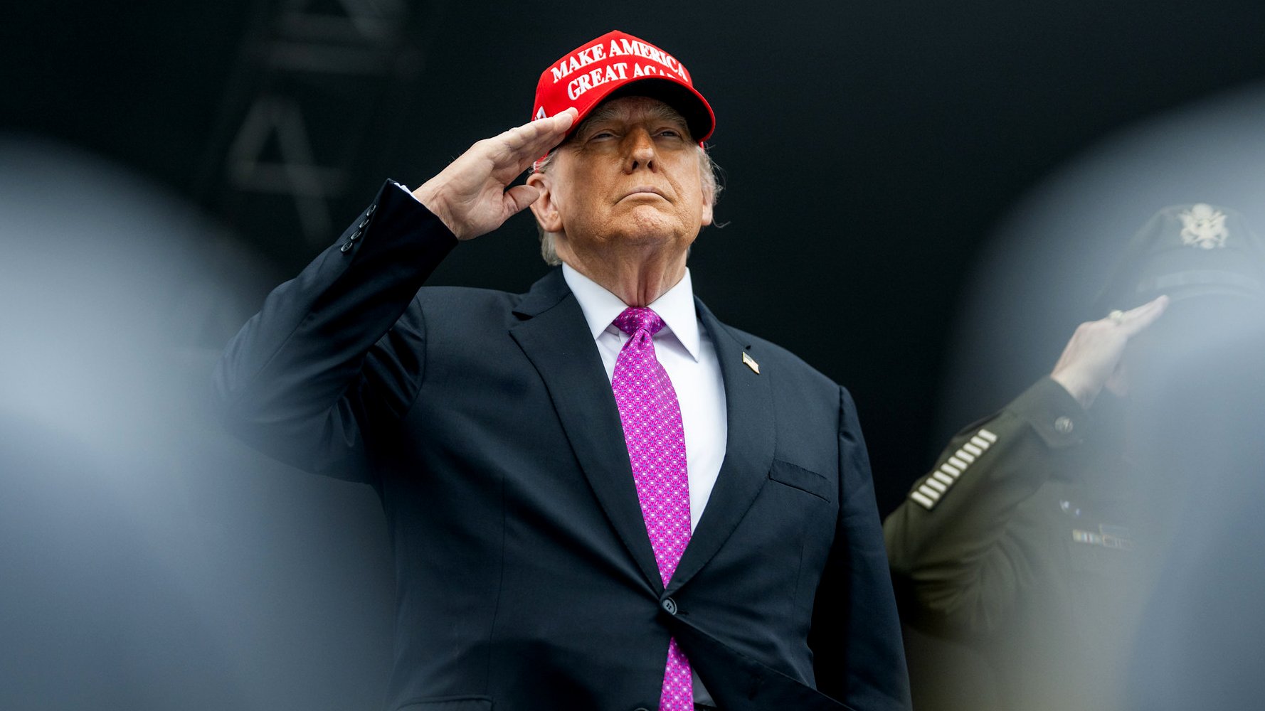 Homme en costume noir, portant une casquette rouge, effectuant un salut.