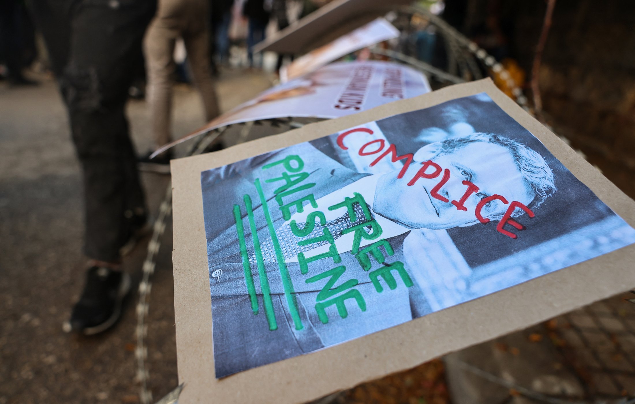 L'immagine mostra un cartello posizionato su una recinzione, con il volto di una persona ritratta in bianco e nero. Sotto l'immagine ci sono delle parole scritte con pennarelli colorati: "COMPLICE" e "FREE PALESTINE". Il contesto sembra essere un evento di protesta, con altri cartelli visibili in background e una folla di persone. L'atmosfera è quella di un'espressione di dissenso e di richiesta di libertà per la Palestina.