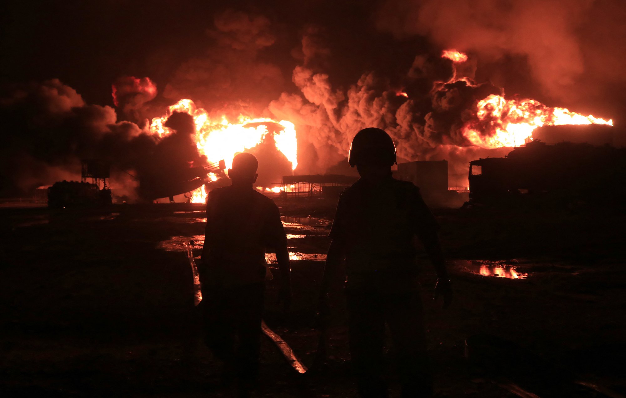 The image depicts two figures walking away from a large fire at night, with bright flames and thick smoke rising into the sky. The scene conveys a sense of urgency and danger.
