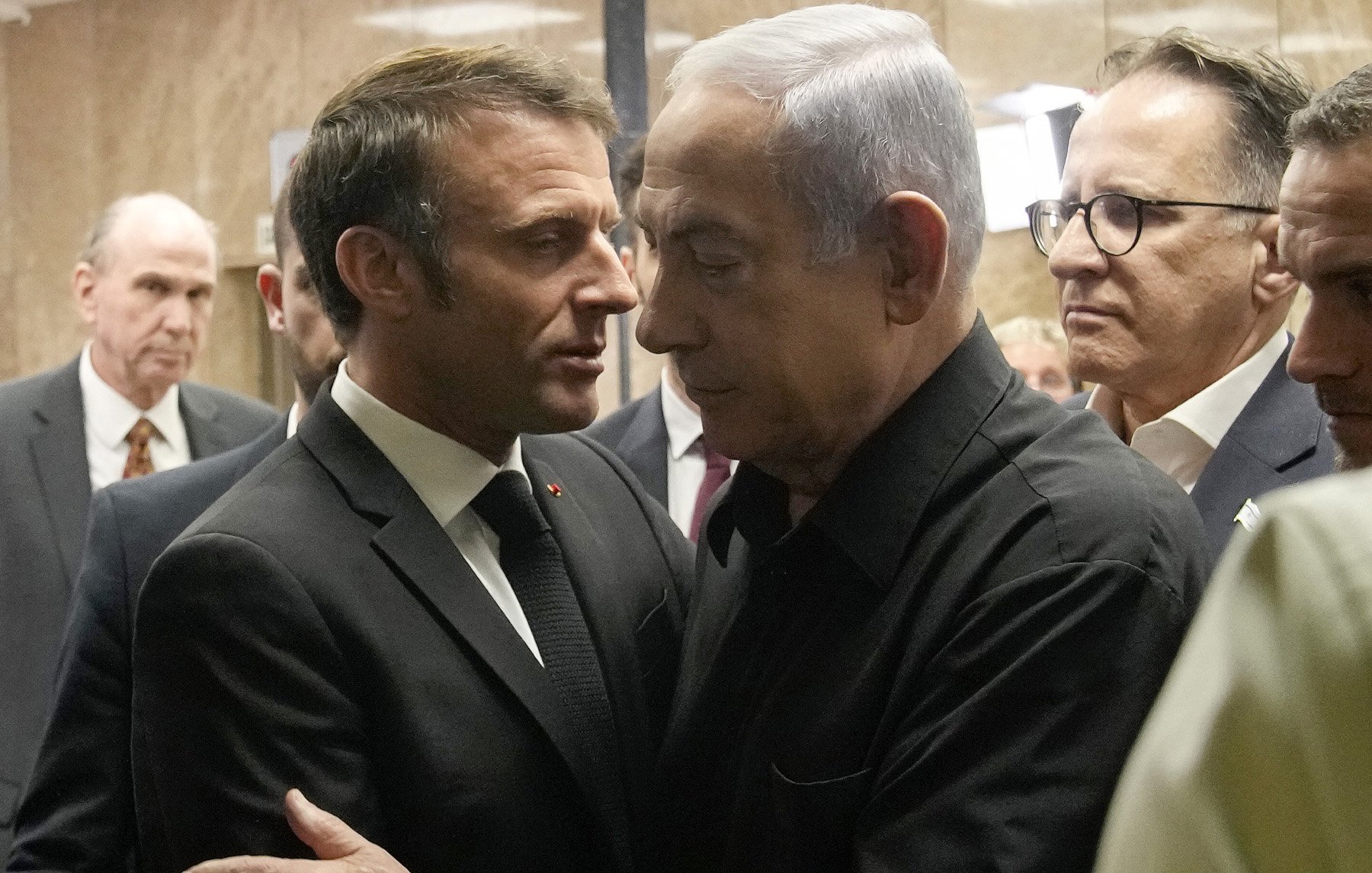 L'image montre deux hommes en discussion proche l'un de l'autre. L'un est en costume noir et le second porte une chemise noire. Ils semblent engageants dans une conversation sérieuse, entourés d'autres personnes en arrière-plan. L'ambiance générale évoque une rencontre formelle ou un événement important.