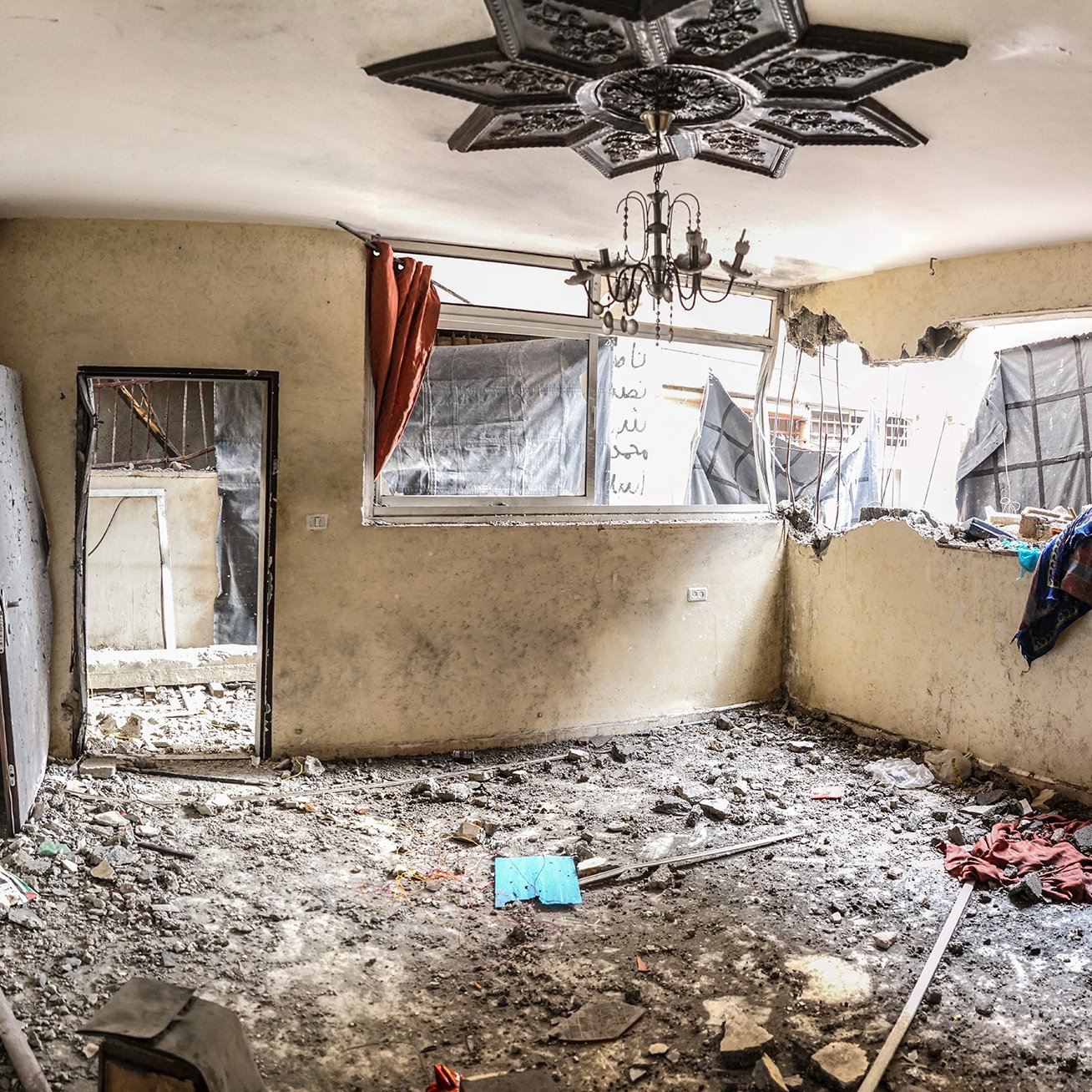 L'image montre une pièce intérieure en mauvais état. Les murs sont abîmés et la peinture est écaillée. Il y a des débris et des gravats sur le sol, ce qui indique un manque d'entretien ou des dommages récents. Une fenêtre est brisée, laissant passer la lumière et révélant des rideaux déchirés. Des vêtements et d'autres objets sont éparpillés, renforçant l'impression de désordre et d'abandon. Un lustre pend du plafond, ajoutant un contraste avec l'état délabré de l'espace. L'ambiance générale semble triste et négligée.